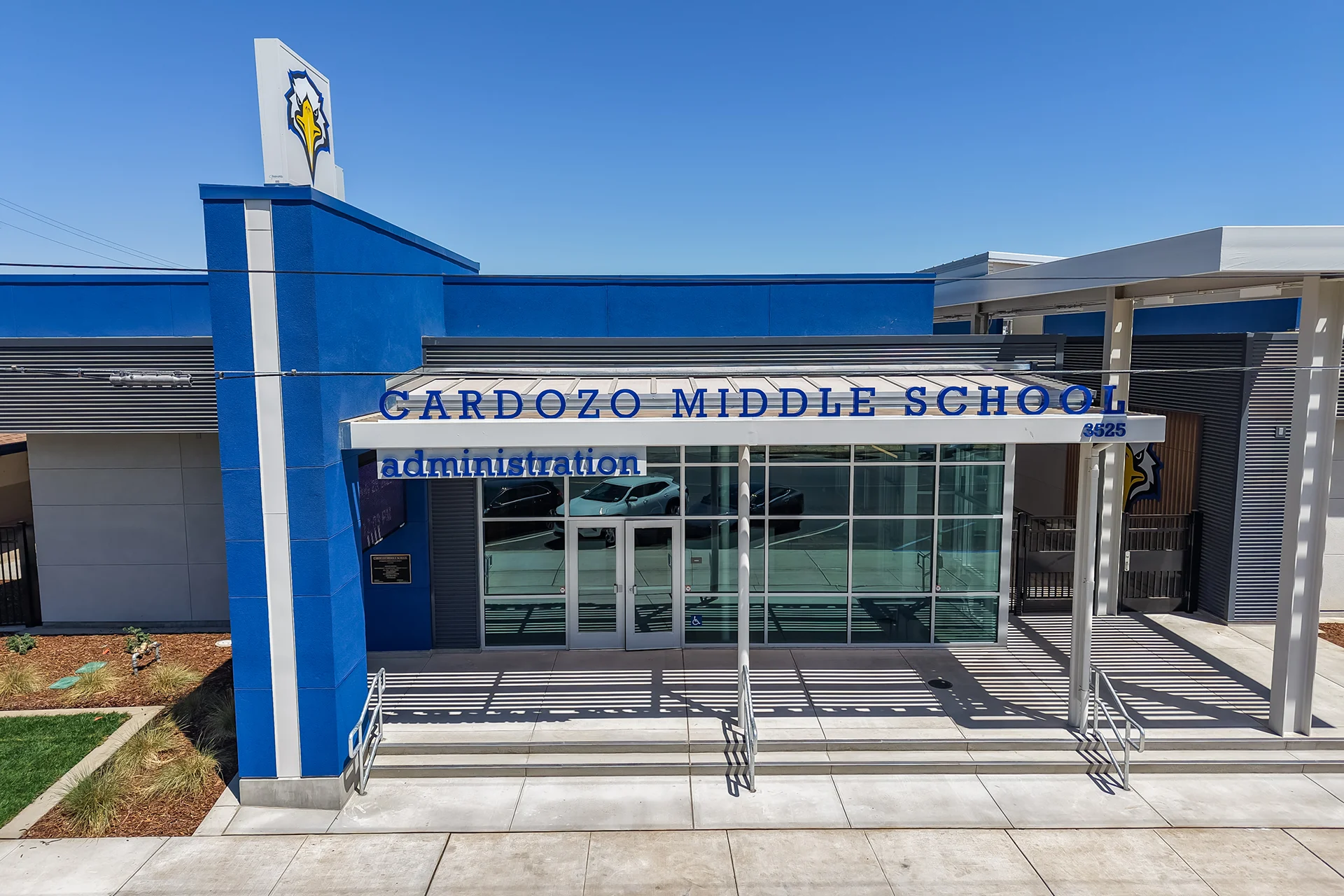 Close-up vertical aerial view of the double glass commercial entry doors at the Cardozo Middle School administration facility. The storefront glass assembly is flanked by structural blue plaster columns and features blue dimensional lettering beneath a custom steel sun-shading canopy canopy design.