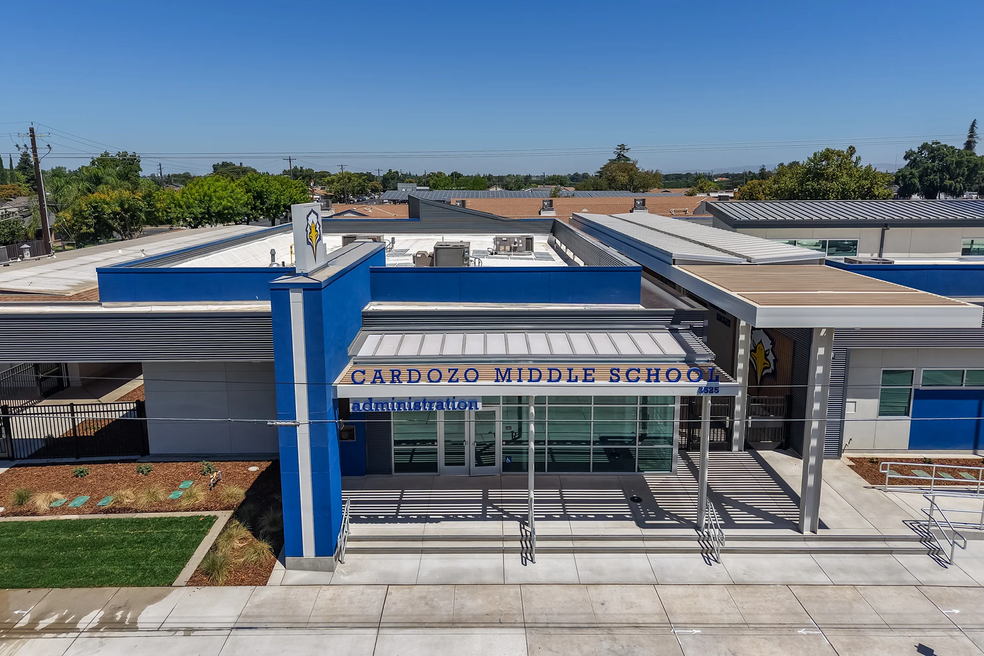 Mid-range drone perspective looking down at the main entryway of the Cardozo Middle School administration wing, highlighting the geometric shadows cast by the custom slatted metal canopy roof over wide concrete steps, integrated steel handrails, a secure perimeter fence line, and fresh green lawn landscaping patches.