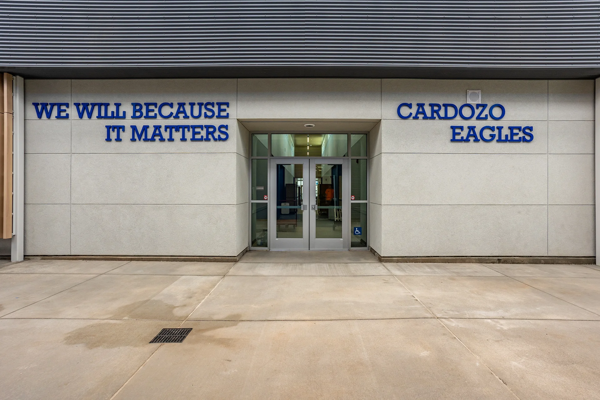Front entrance exterior view of a school campus building, displaying double glass entry doors encased in aluminum storefront framing. The smooth architectural concrete plaster facade features dimensional blue metallic lettering stating We Will Because It Matters and Cardozo Eagles under corrugated metal wall cladding.