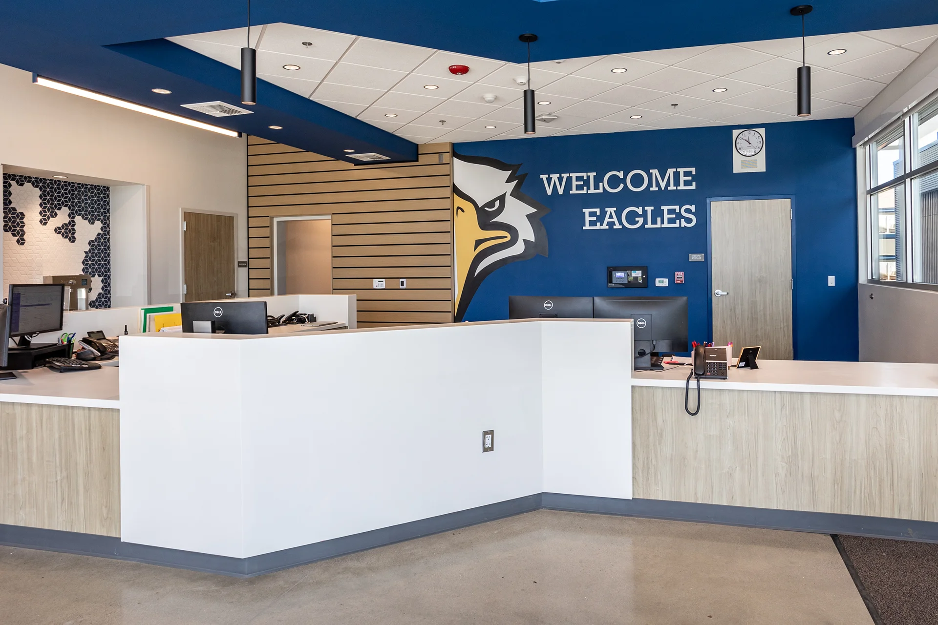 Front view of the main institutional office reception desk and secure waiting area, showcasing modern built-in workspaces behind a white privacy partition wall. The background displays a prominent blue focal wall with dimensional block lettering, light oak laminate doors, and a decorative blue-and-white hexagonal tile mosaic backsplash.