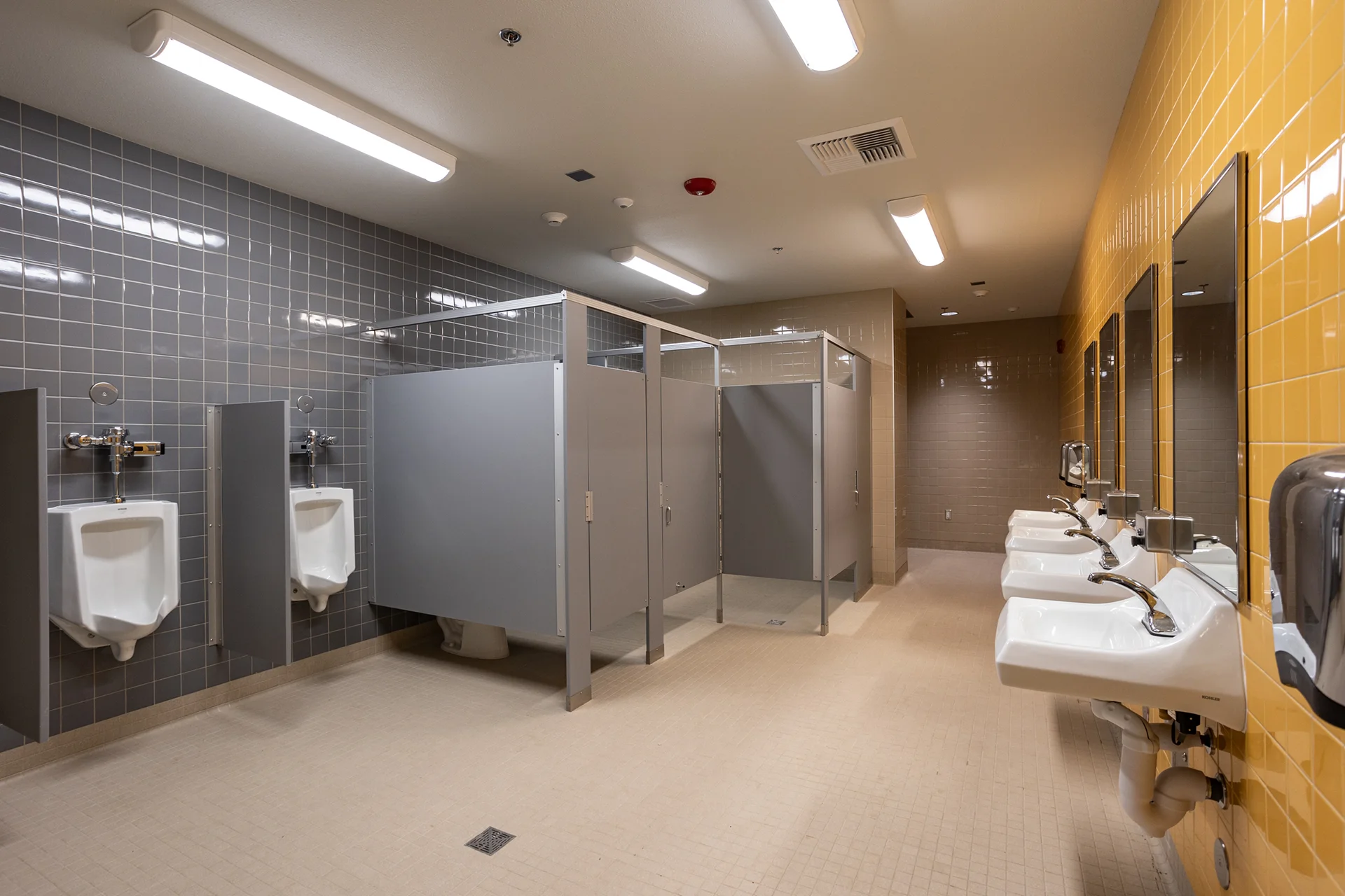 Institutional student restroom constructed by CT Brayson & Sons, displaying wall-hung vitreous china urinals separated by grey laminate privacy screens against a dark grey grid tile wall. The facility includes automatic flush valves, centralized floor drains, a row of sinks, and contrasting yellow tiled accent walls.