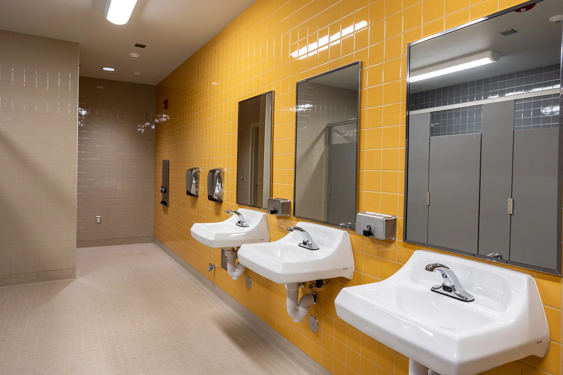 Wide view of an institutional school washroom vanity wall clad in glossy yellow tile squares. A series of white porcelain washbasins with automatic motion-activated chrome faucets line the wall alongside matching chrome soap dispensers, trash receptacles, and vanity mirrors.