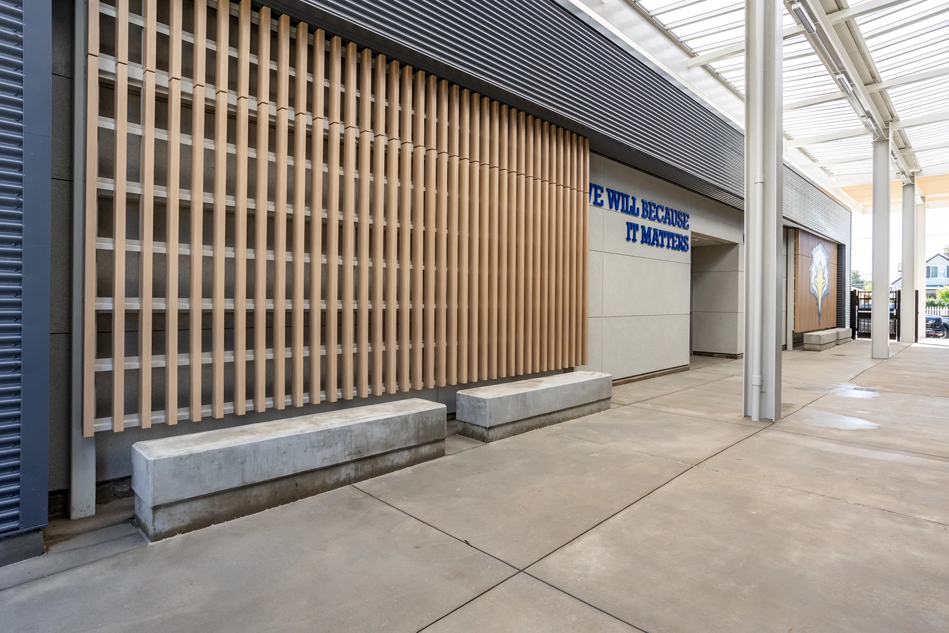 Campus exterior walkway corridor showing cast concrete structural benches positioned in front of a decorative vertical wood slat sunscreen façade element. The outdoor space features a concrete sidewalk plaza under a protective metal canopy roof with high steel supporting columns.