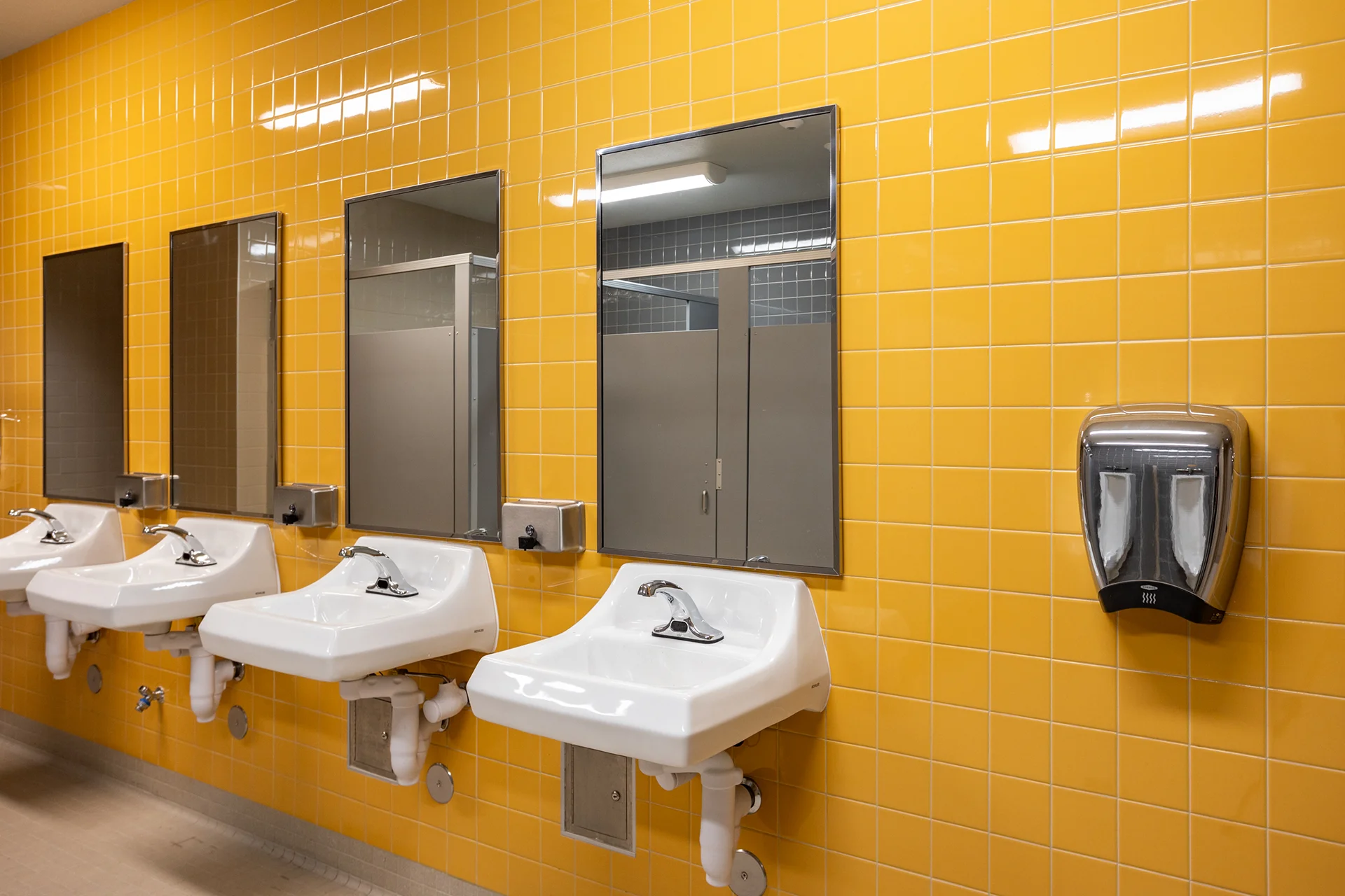 Commercial washroom facility interior showing white porcelain drop-in basins fitted with automatic chrome faucets against a glossy yellow tiled wall. The layout features individual rectangular vanity mirrors and a wall-mounted stainless steel automated hand dryer.