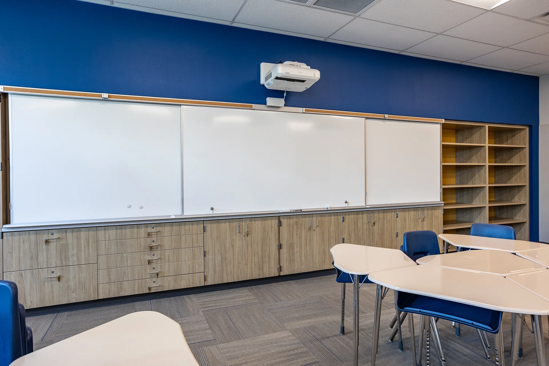 Close-up commercial millwork detail inside a school facility showing custom wood-grain laminate base cabinetry with lockable storage drawers. The storage unit is topped with a neutral laminate countertop below a continuous sliding whiteboard track system and a built-in open-shelf book casing.