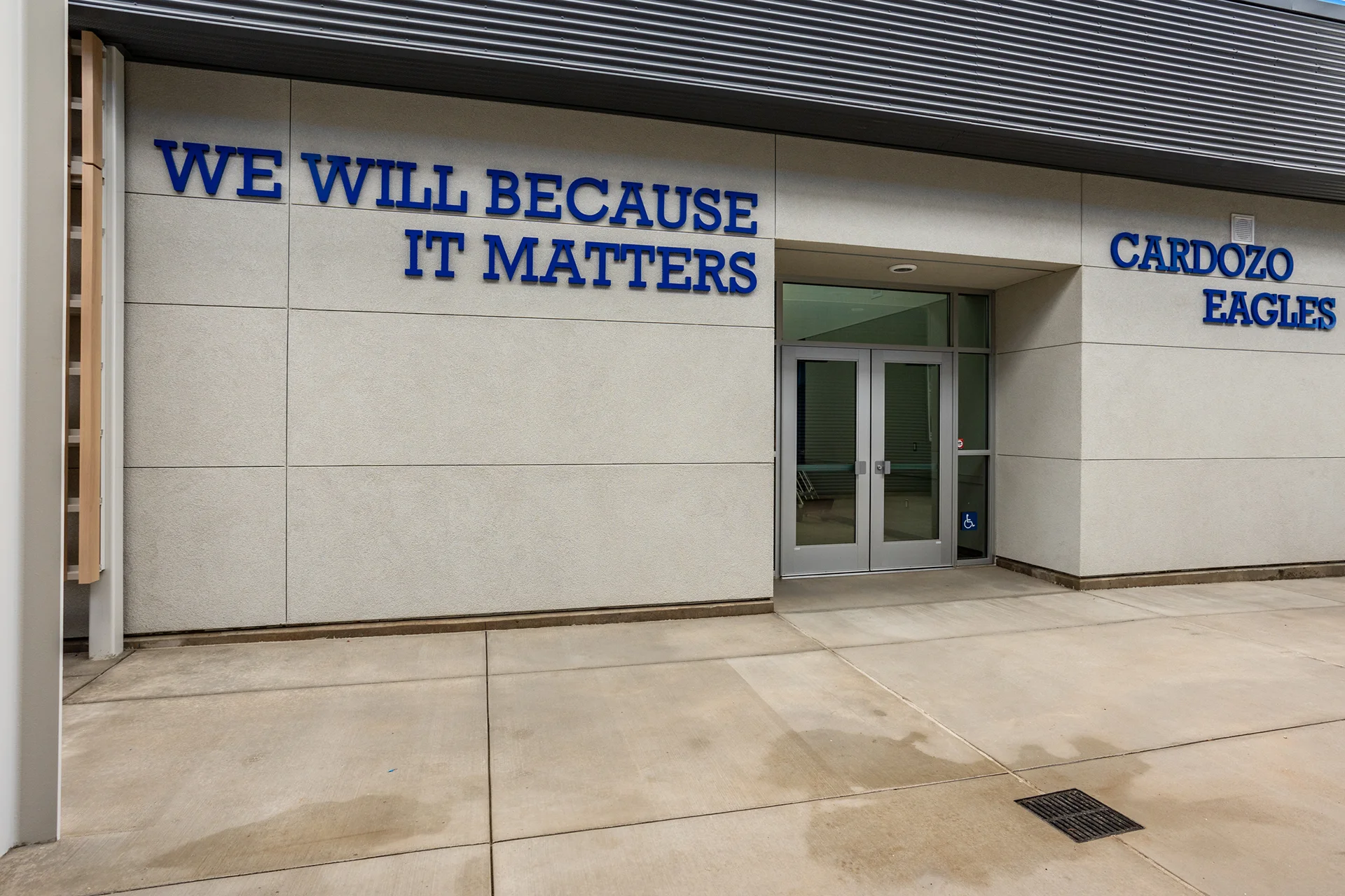 Close-up structural exterior perspective of a school campus facility entrance, highlighting a pair of clear glass commercial storefront entry doors with integrated push bar exit hardware, an ADA accessibility door button, smooth concrete wall panels, and an overhead corrugated steel awning profile.
