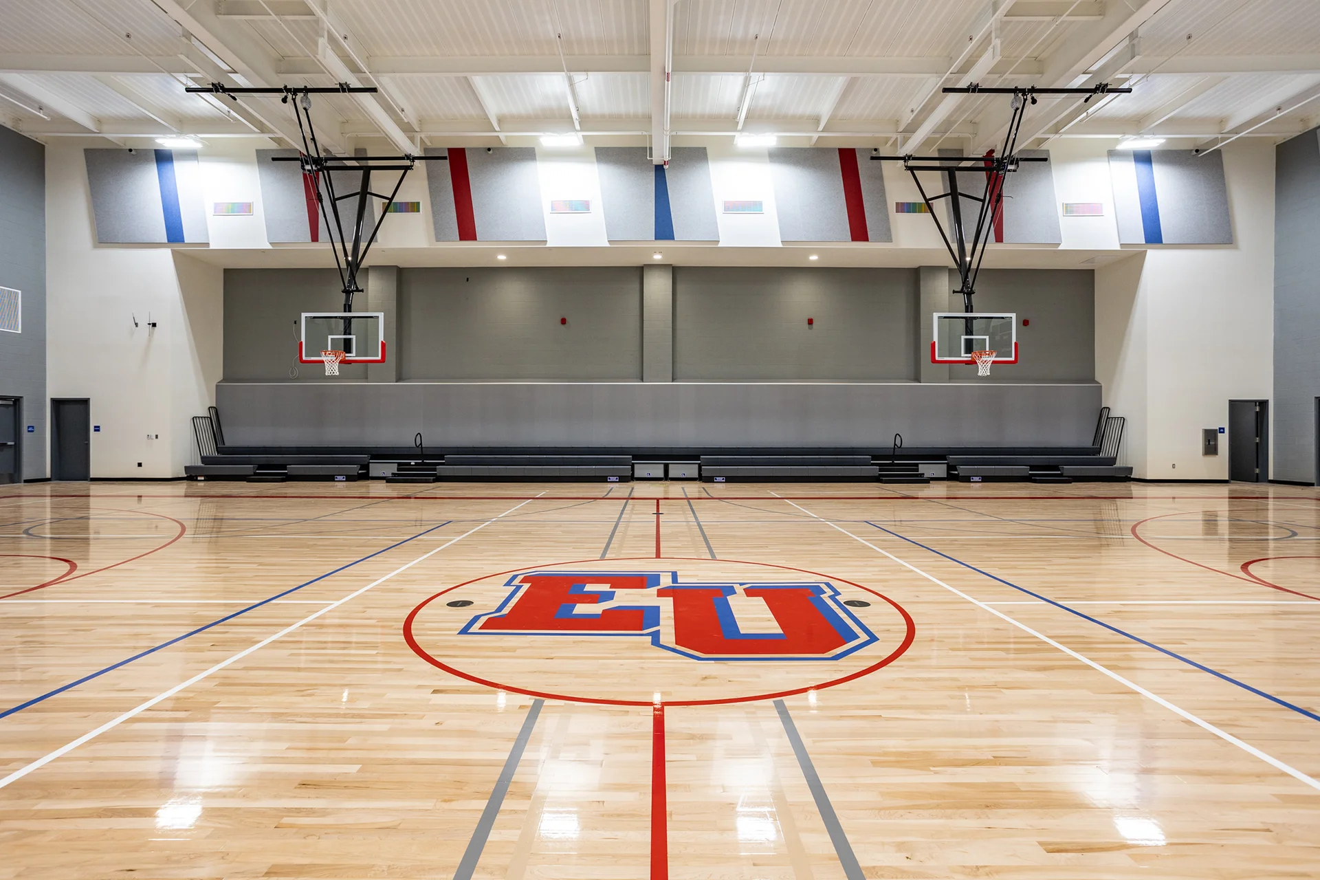 Detailed indoor athletics center highlighting a high-gloss polished hardwood basketball court with red and blue painted lines, ceiling-hung retractable acrylic backboards, gray wall bleachers, and decorative acoustic wall baffling.