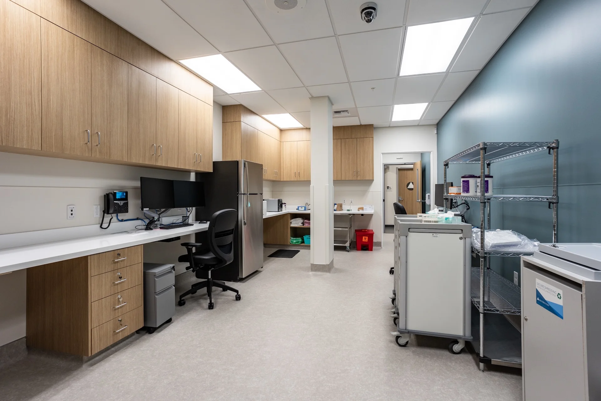 Wide perspective of a medical facility's central prep station, complete with wrap-around wood grain cabinetry, seamless anti-microbial countertops, dual monitors, a full-sized stainless steel medical refrigerator, wire storage shelving units, and rolling supply carts on resilient vinyl flooring.
