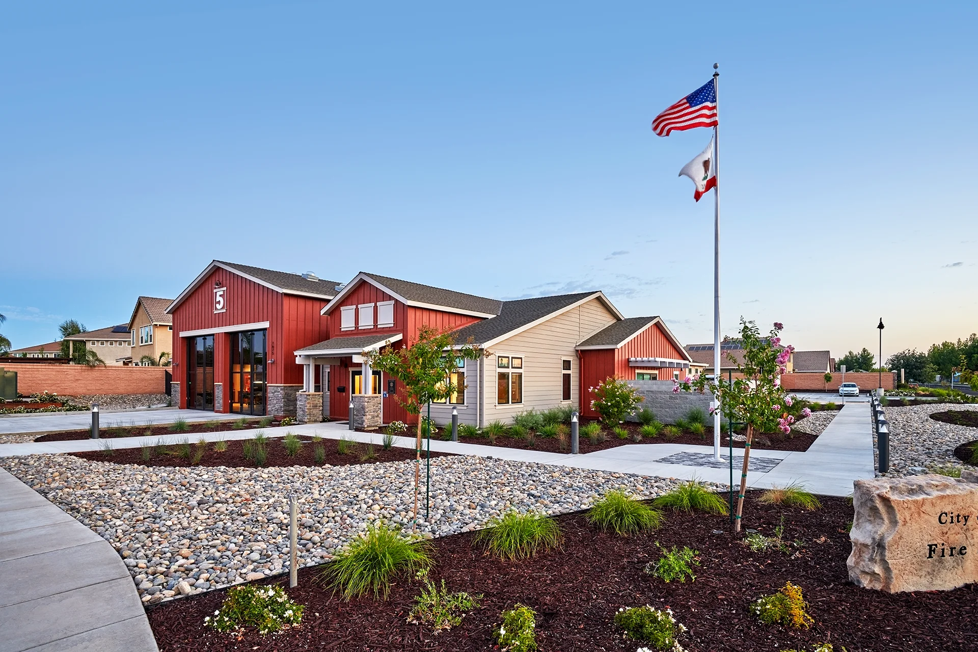 Wide-angle evening shot of the newly constructed fire station in Manteca, California, highlighting the illuminated glass garage bay doors, manicured mulch beds, concrete walkways, and perimeter security lighting.
