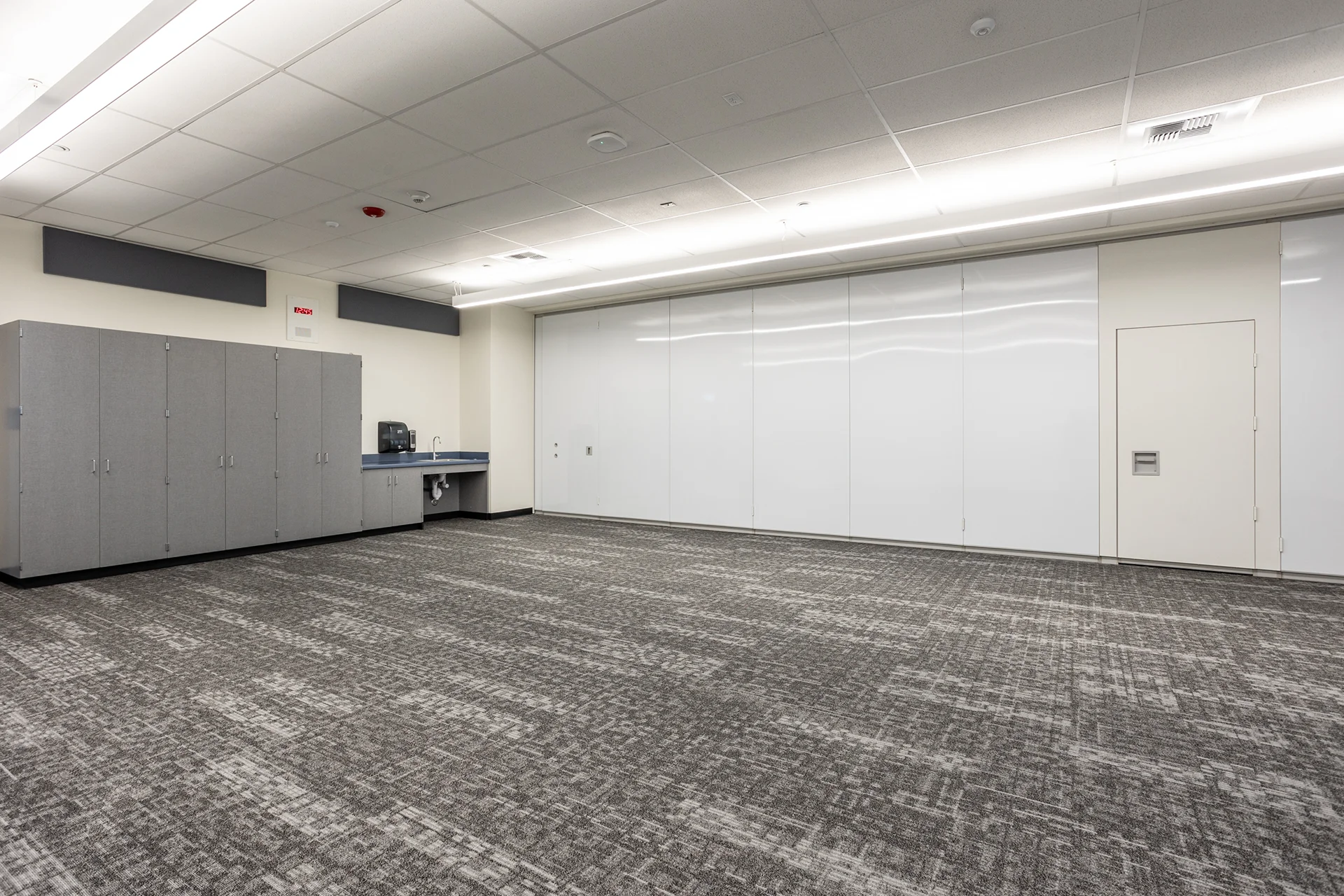 Academic learning space showing a floor-to-ceiling white dry-erase whiteboard wall, architectural recessed linear strip lights, gray commercial carpet tile, and built-in laminate cabinetry with an active utility sink.