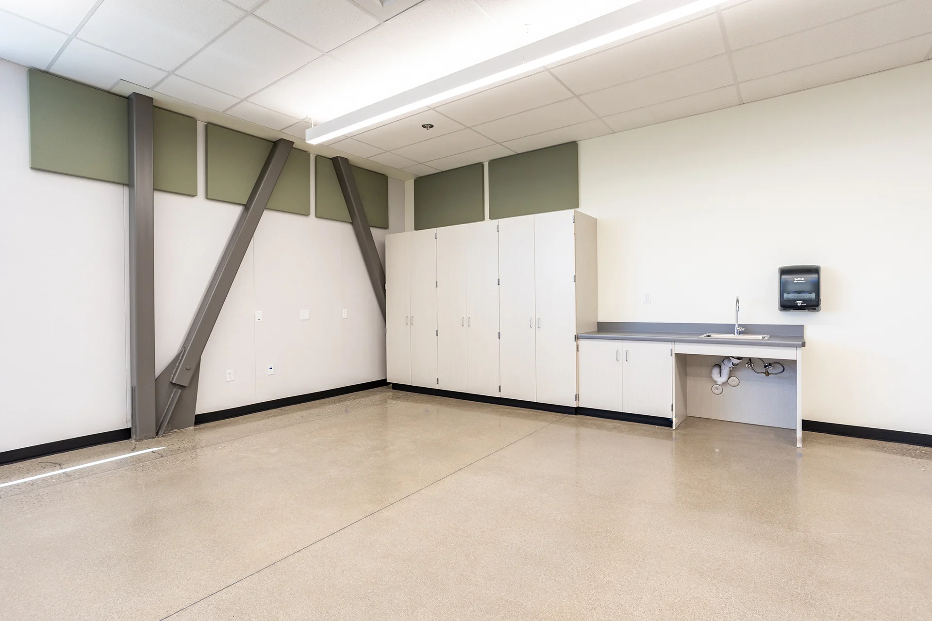 Wide angle interior perspective of an institutional room showing built-in light maple laminate cabinetry, an ADA-compliant cutout utility counter with a stainless steel sink, heavy-duty industrial structural steel cross-bracing along the white wall, and a high-gloss sealed concrete floor.