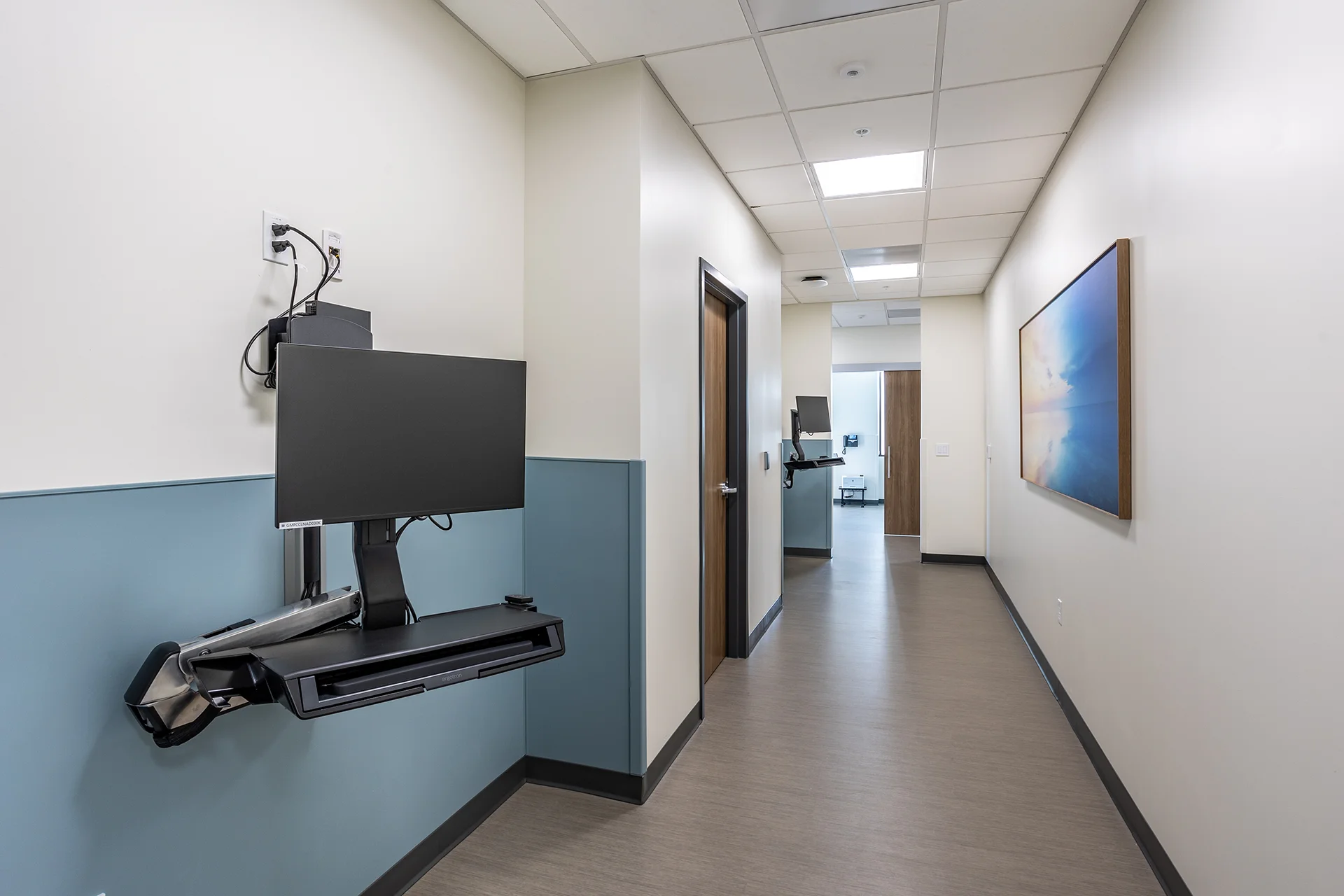Main hallway inside a medical facility in Manteca displaying an Ergotron wall-mounted sit-stand computer station with an integrated keyboard tray, teal-painted wainscoting, solid core wood doors with commercial hardware, and flush-mount LED corridor lighting.