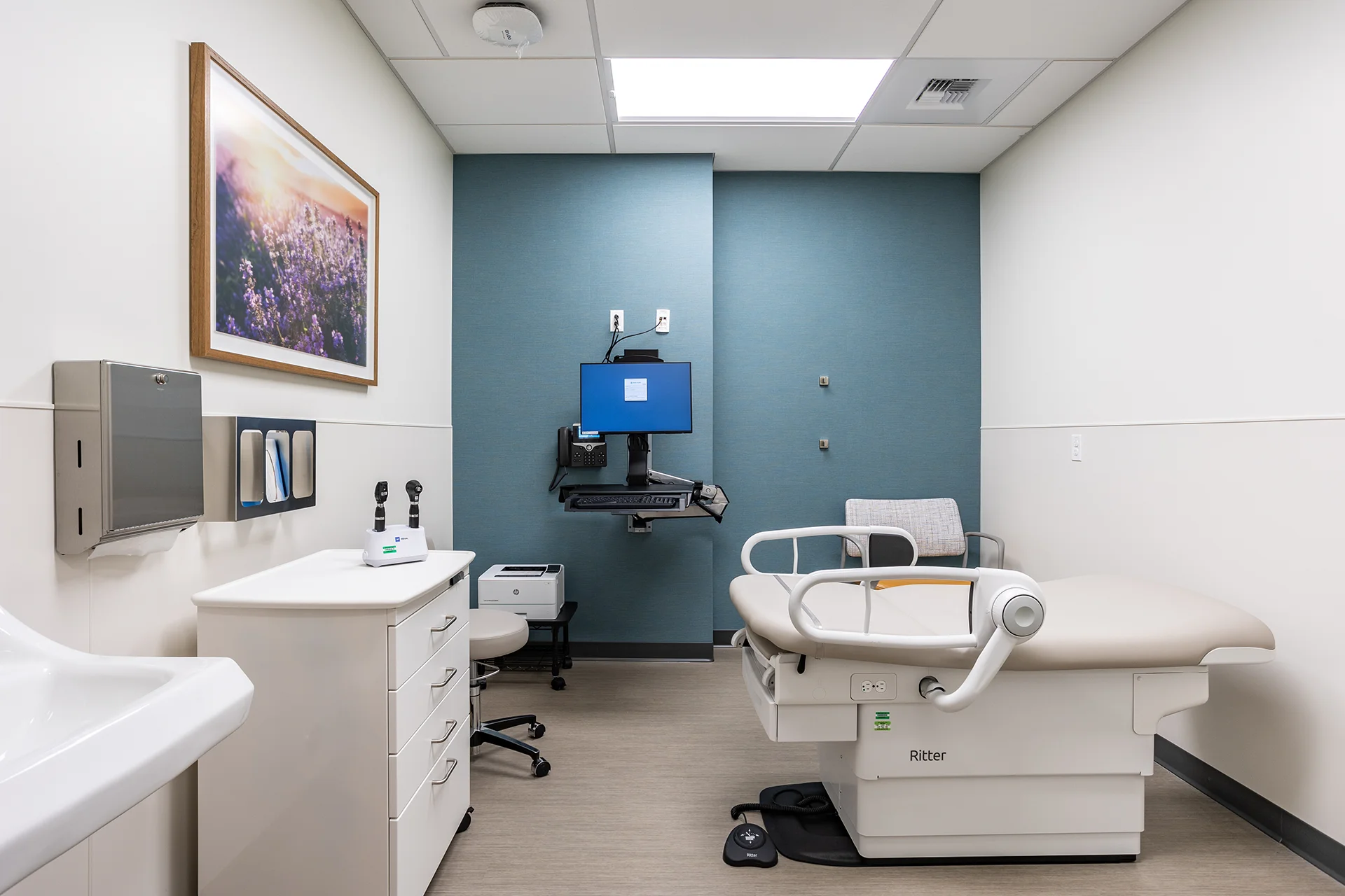 Modern medical examination room in Manteca, California, featuring a white Ritter adjustable bariatric exam table, wall-mounted diagnostic station with flat-screen computer monitor, custom multi-drawer white melamine storage cabinets, and a teal blue accent wall.