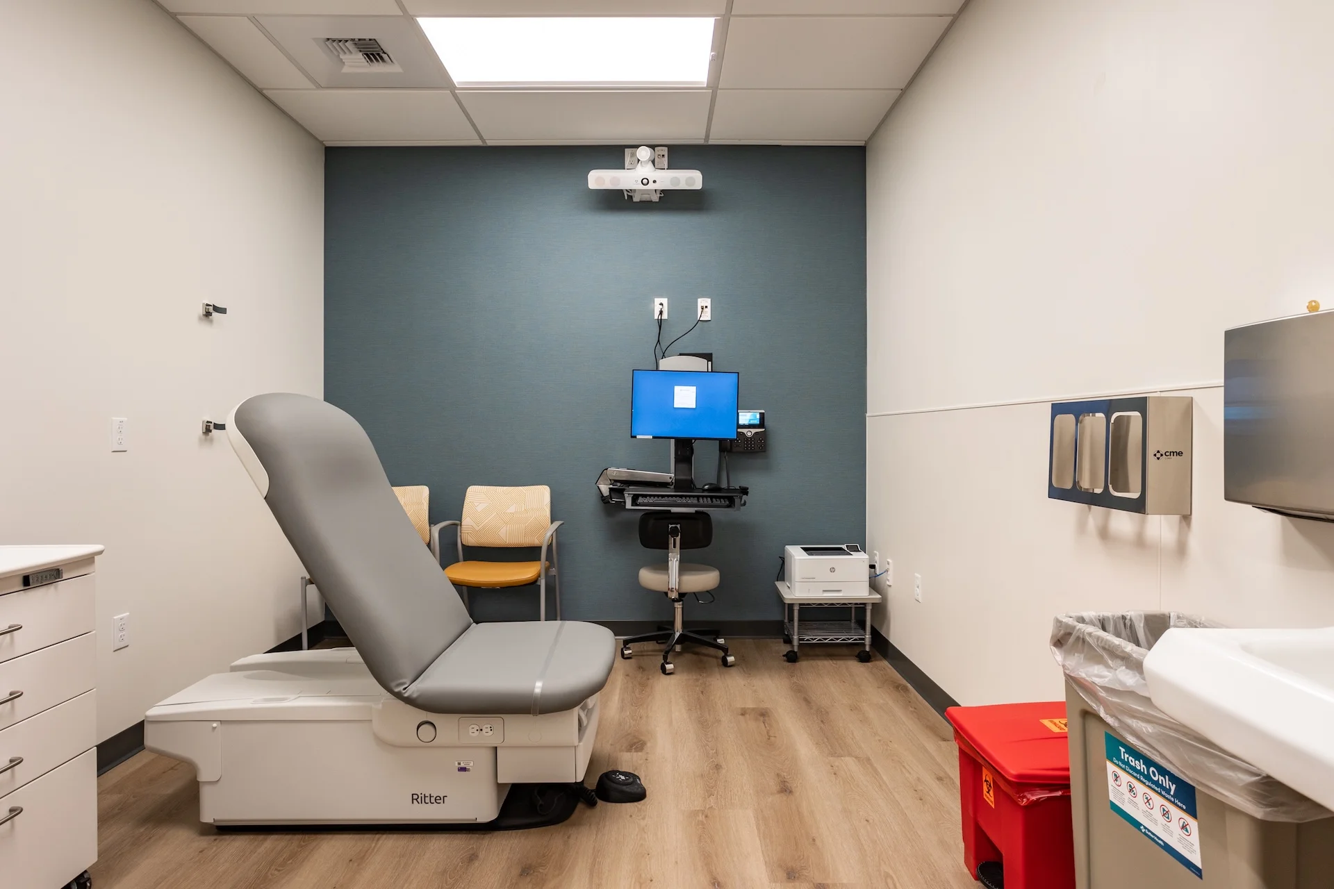 Standard clinical exam room interior featuring a dual-tone adjustable examination chair, wall-mounted computer cart workstation, wood-look luxury vinyl plank flooring, neutral painted drywall, a hands-free handwashing sink station, and specialized medical waste disposal units.