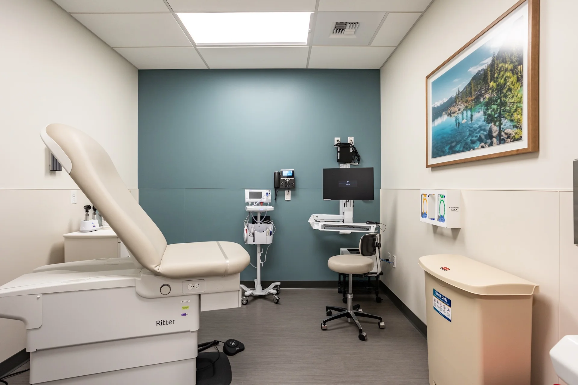 Dedicated medical examination room featuring an adjustable electric patient chair, a rolling physician's stool, an integrated wall charting station with a flatscreen monitor, automated handwashing facilities, and scenic landscape wall decoration.