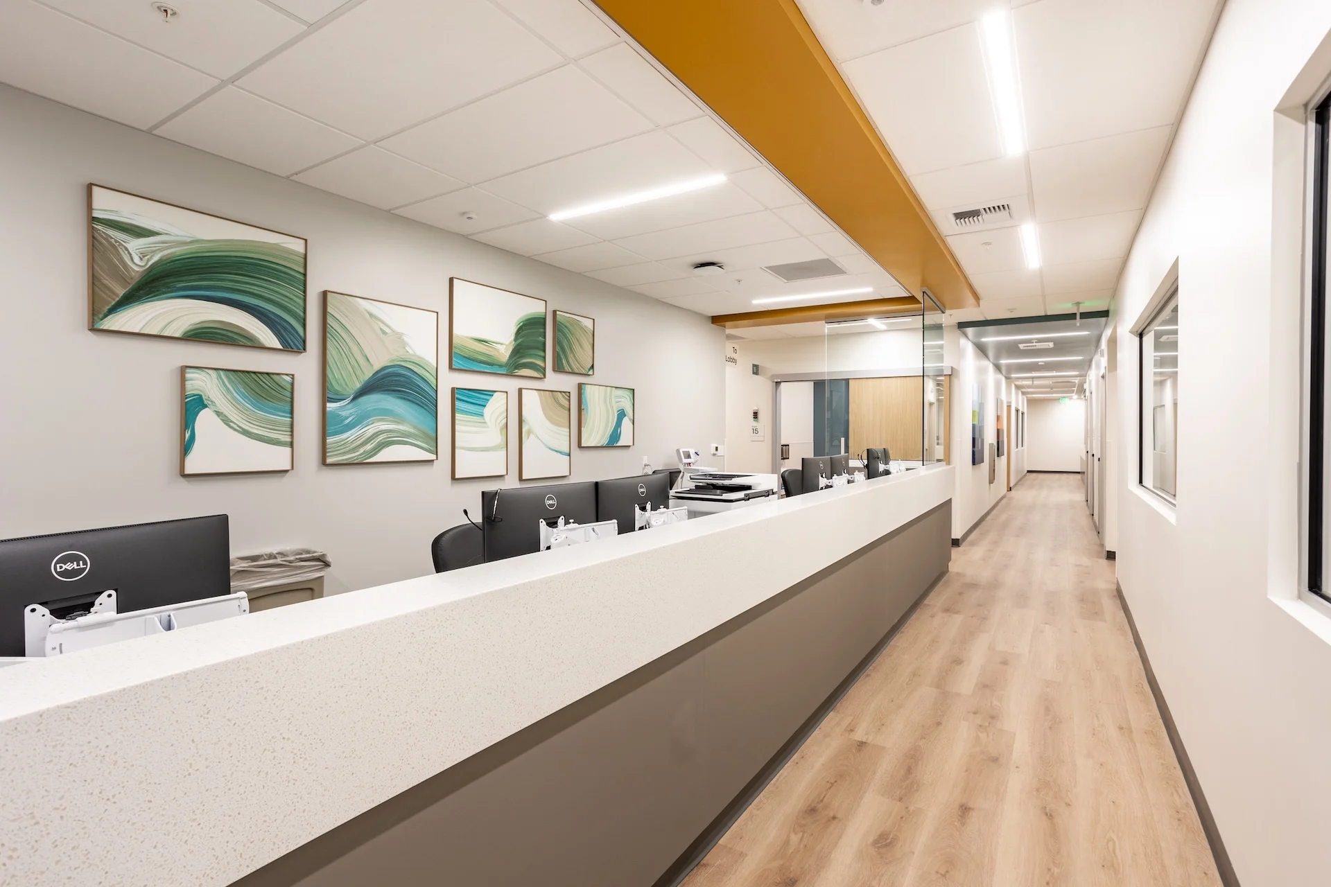 Architectural perspective of a healthcare facility check-in station showcasing an expansive solid-surface quartz counter, custom wood-framed canvas art panels, a drop-ceiling accent painted in a warm mustard hue, and low-profile LED linear light troffers highlighting a smooth commercial-grade plank hallway.