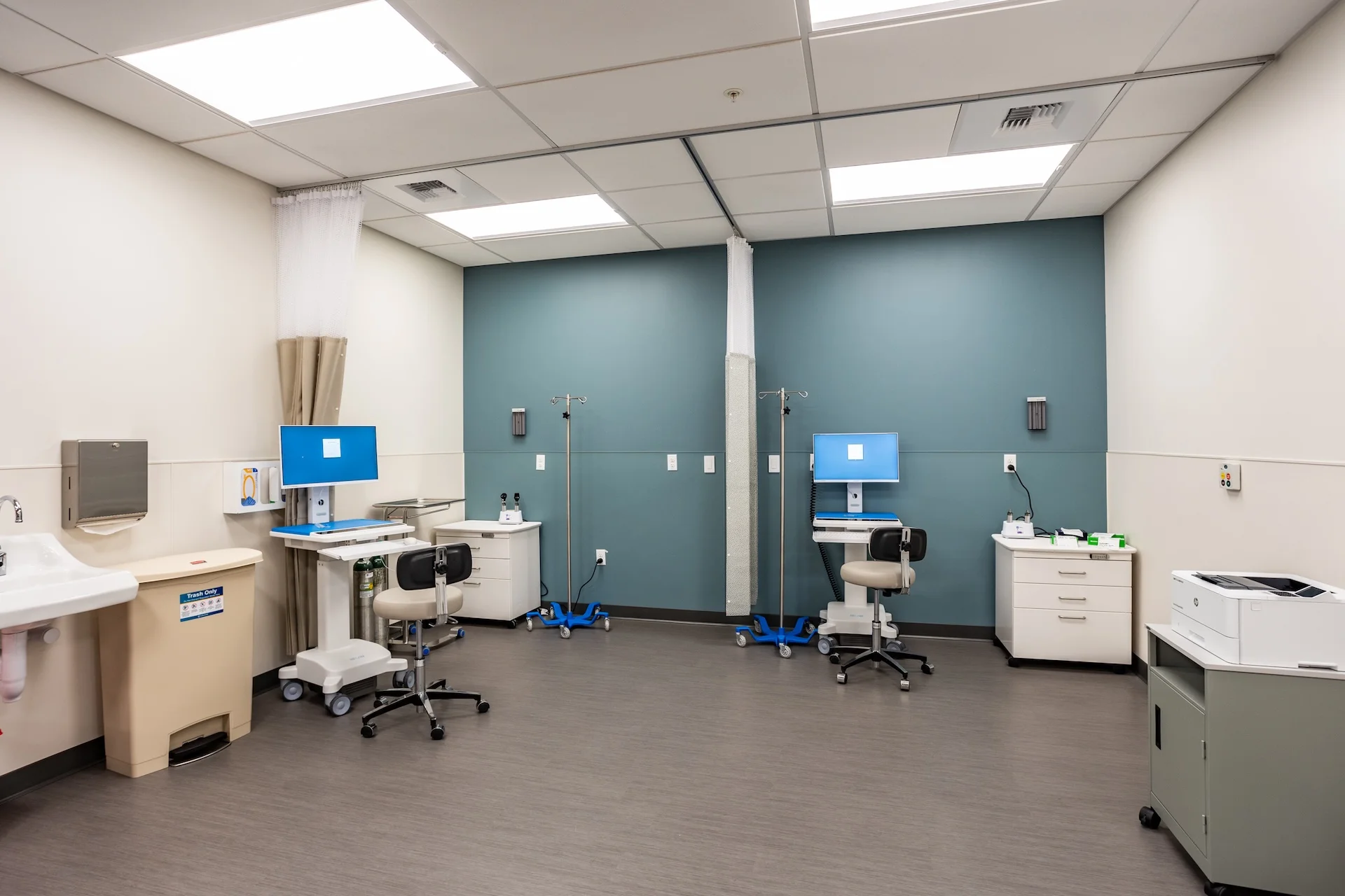 Interior view of a clinical healthcare space configured with multiple patient recovery bays separated by privacy curtains, showing custom white laminate medical cabinetry, stainless steel sinks, integrated medical gases and data ports, rolling medical stools, and sophisticated computer charting stations under linear recessed LED lighting.