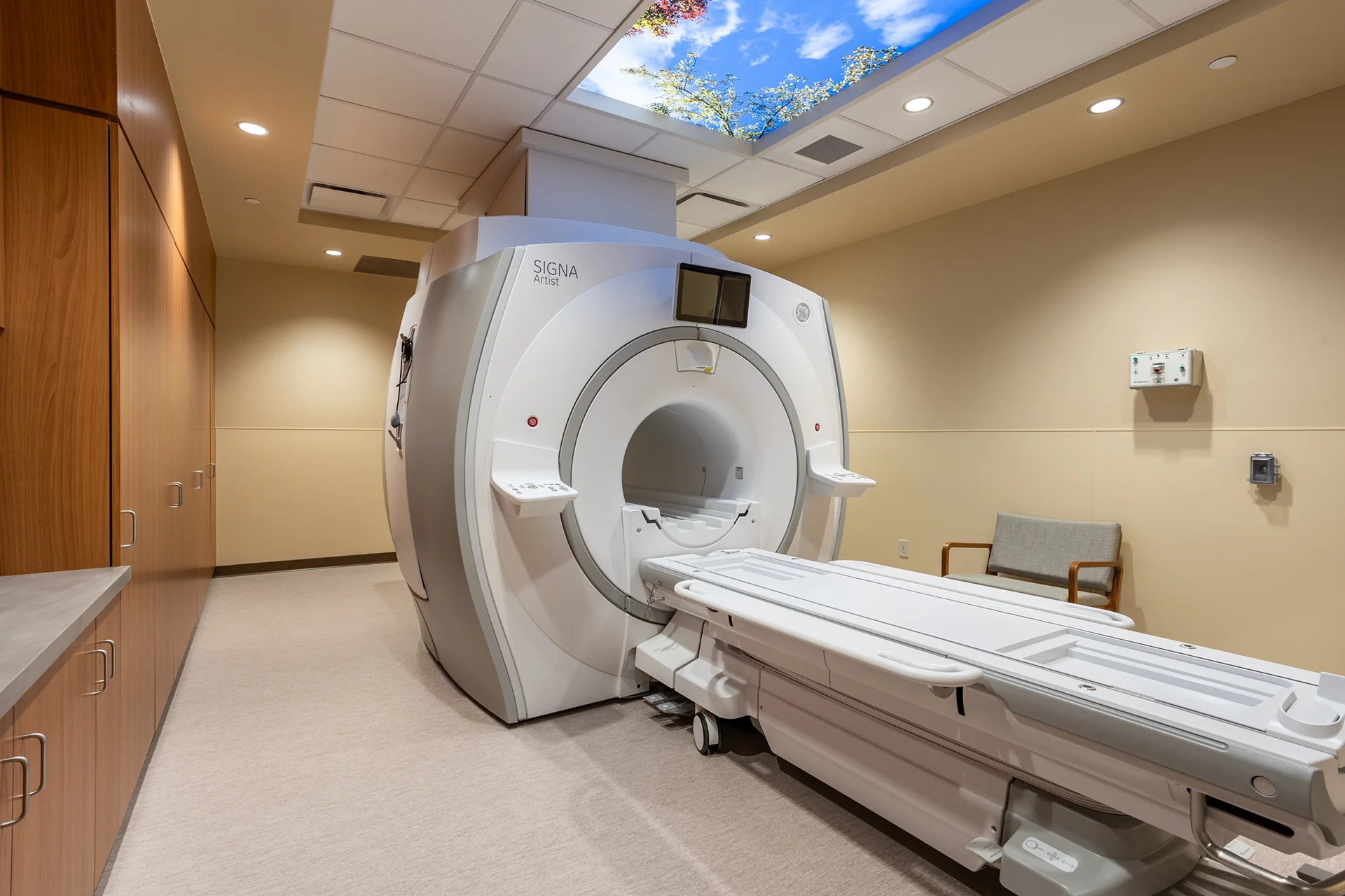 Diagnostic suite interior showing a large white magnetic resonance imaging machine with a patient transport table, custom floor-to-ceiling wood storage millwork, and a backlit nature graphic ceiling fixture.
