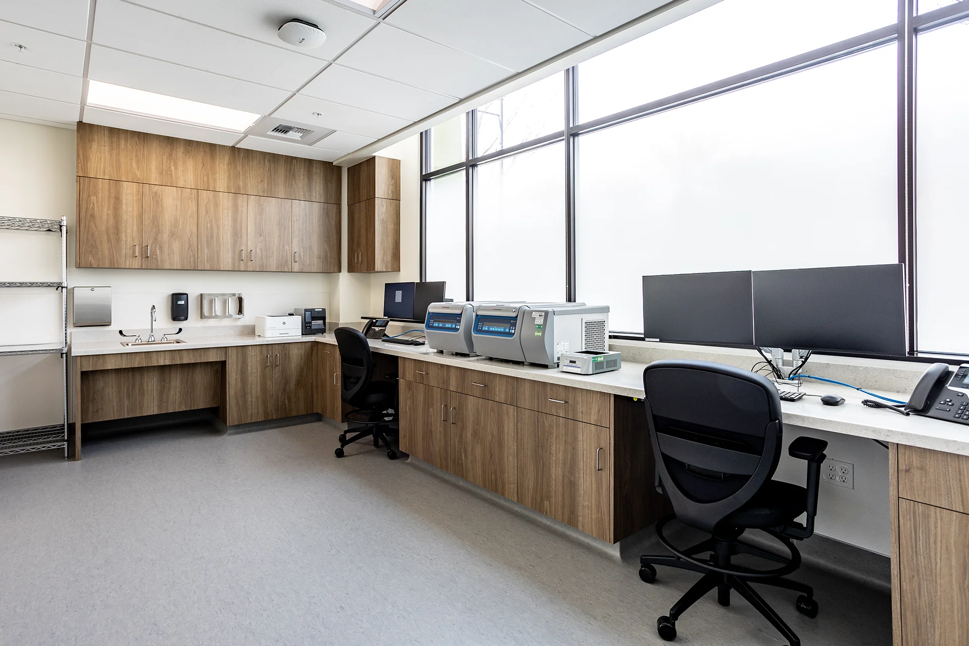 Wide-angle view of a medical clinic laboratory in Central California, displaying extensive walnut-grain base cabinets, an integrated handwashing alcove, rolling ergonomic lab chairs at a computer desk, and dual Thermo Scientific specimen centrifuges under bright LED ceiling panels.