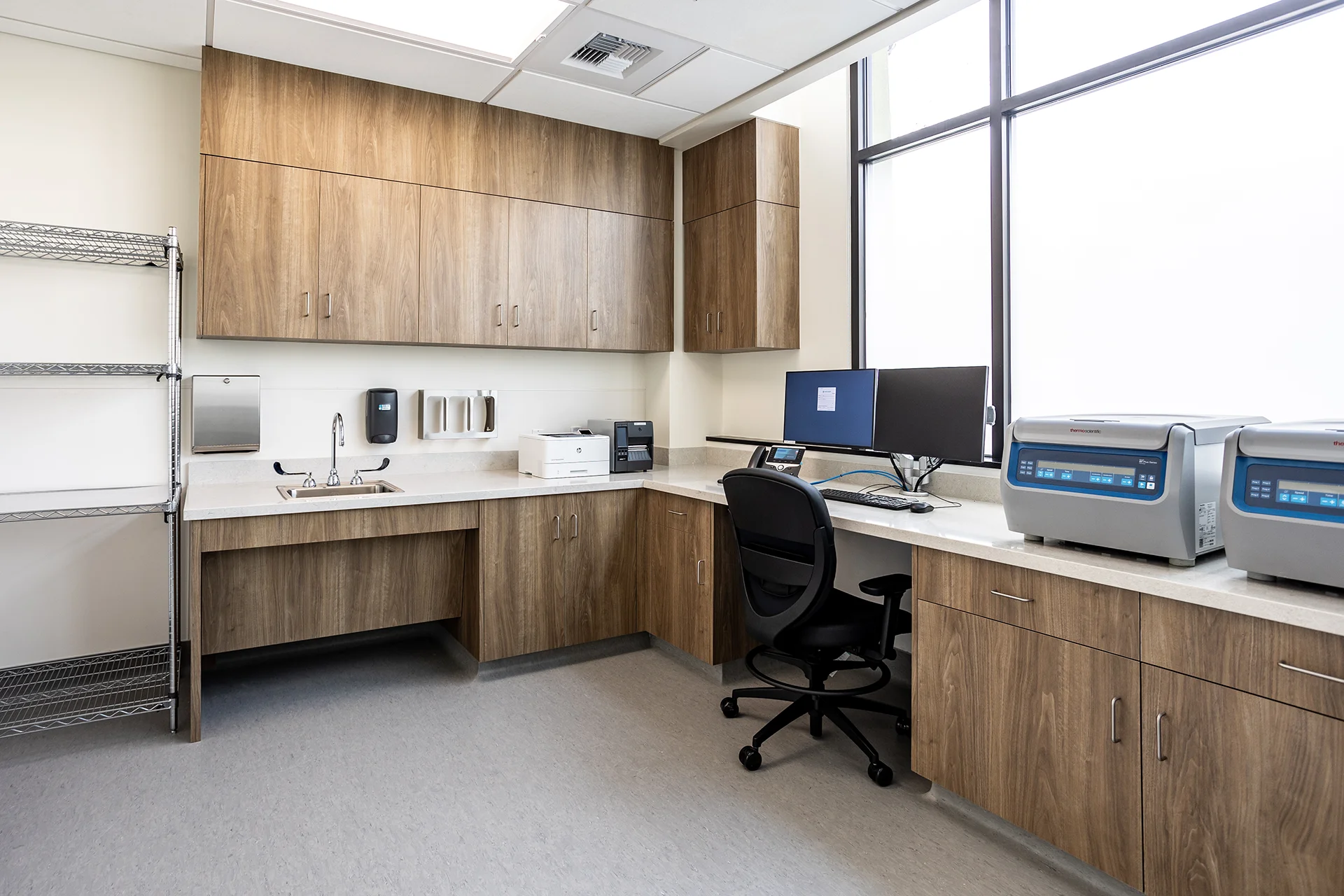 Laboratory testing environment in Manteca highlighting dual Thermo Scientific clinical centrifuges on a solid-surface workspace, built-in wood-grain storage cabinets, a stainless steel handwashing sink, and large windows with privacy film.