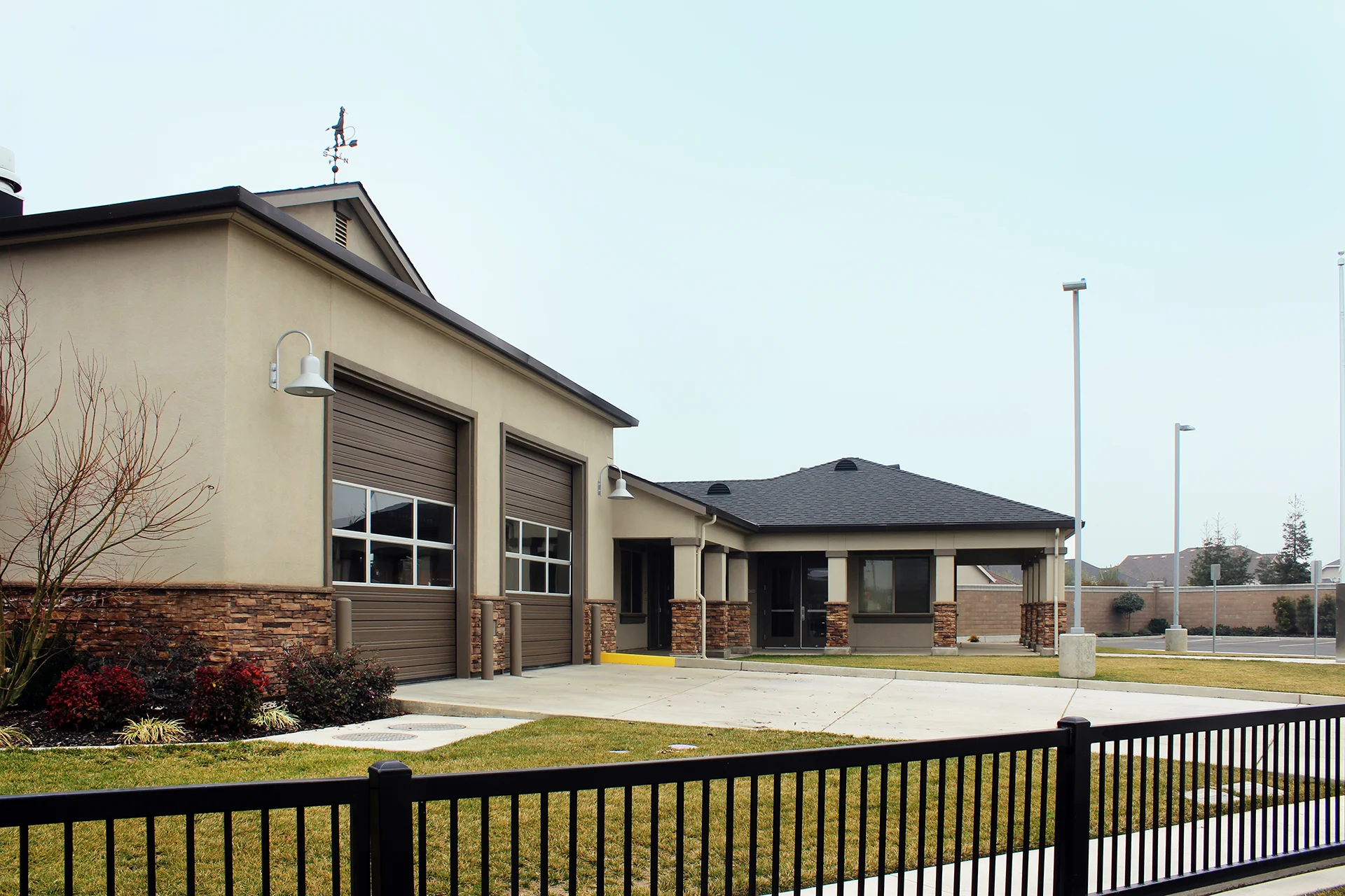 Angled architectural detail of the station's response garage bays, showcasing decorative cultured stone wainscoting along the foundation wall, modern dark brown sectional roll-up doors with glass window rows, and silver gooseneck light fixtures.