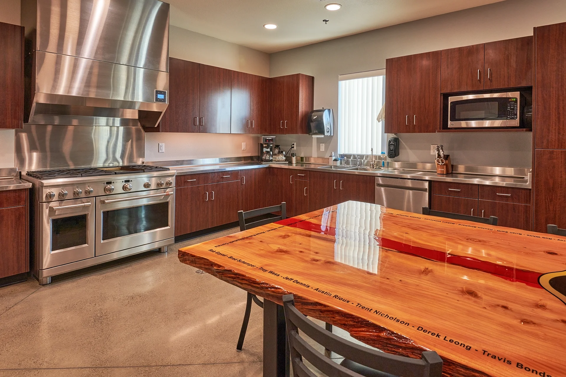 Detailed view of the firehouse kitchen construction, emphasizing the high-gloss epoxy finish on a custom wood slab table, an industrial stainless steel range hood, and integrated kitchen appliances.