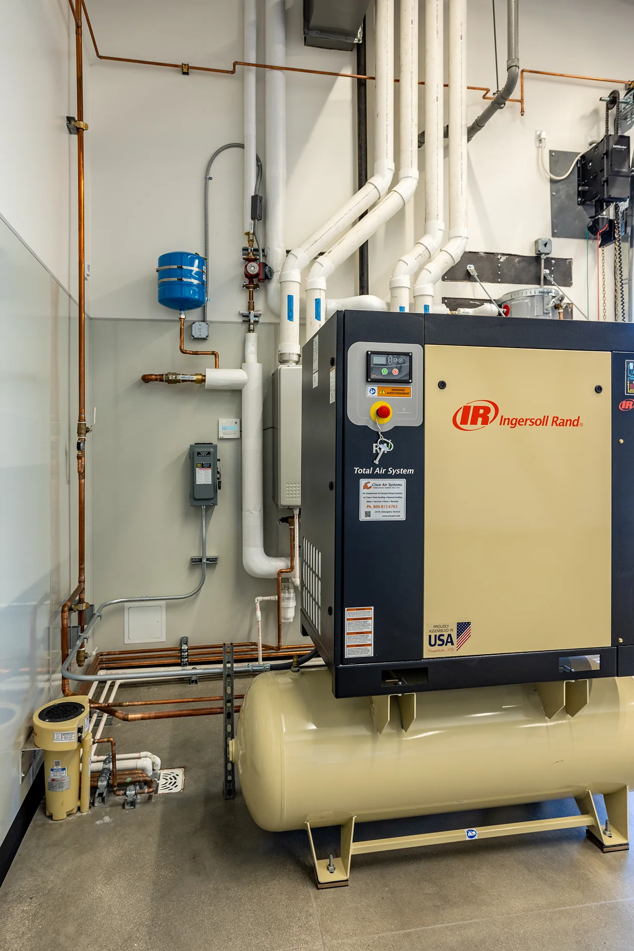 Vertex view of a high-capacity industrial air compressor system mounted on a horizontal storage tank, featuring complex copper plumbing lines, insulated overhead utility pipes, and electrical disconnect boxes designed for facility maintenance.