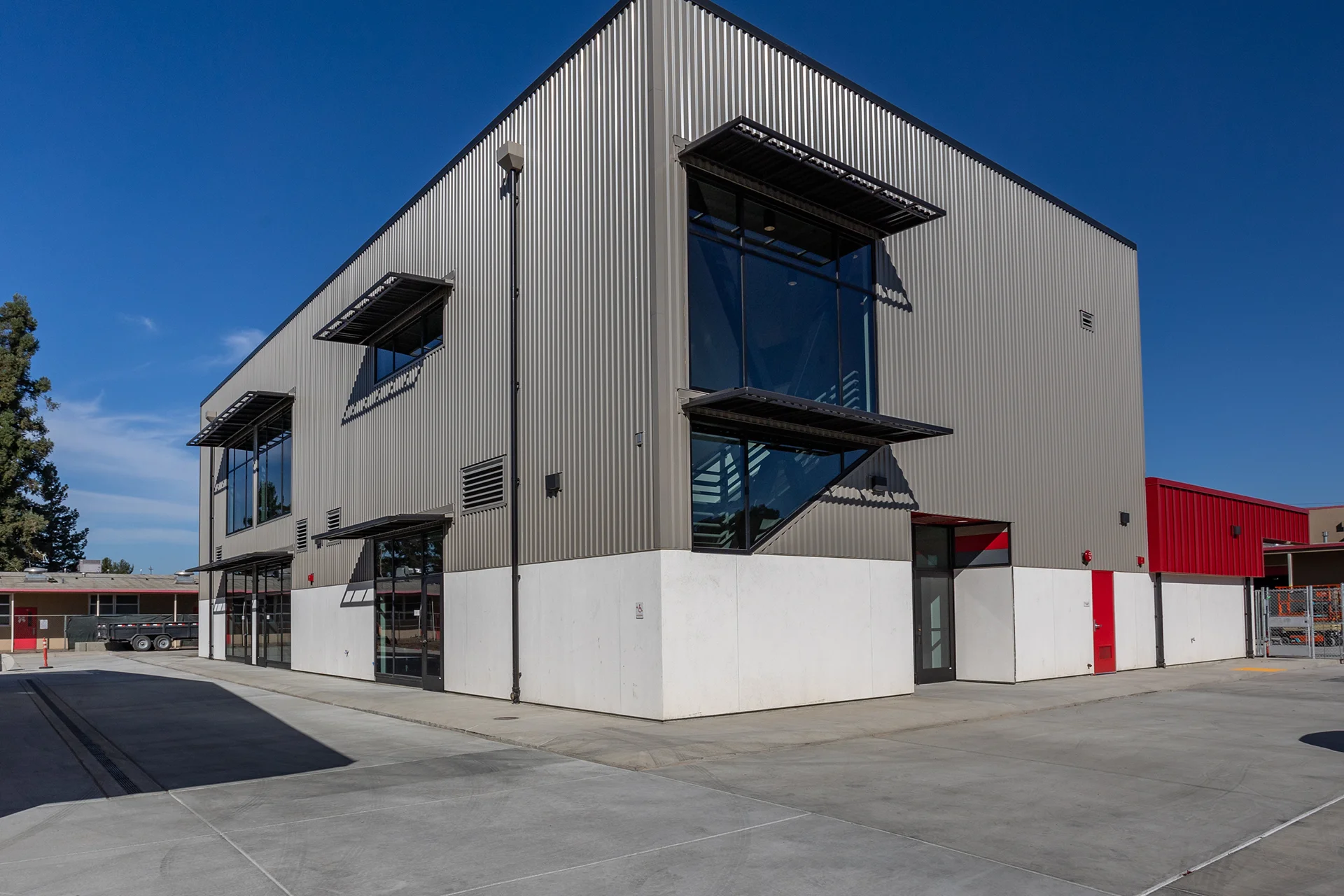 Exterior perspective of a multi-story modern industrial structure engineered with vertical corrugated steel siding, a stark white concrete foundation base wall, multi-lite commercial windows fitted with black metal louvered sunshades, and a clean concrete apron.