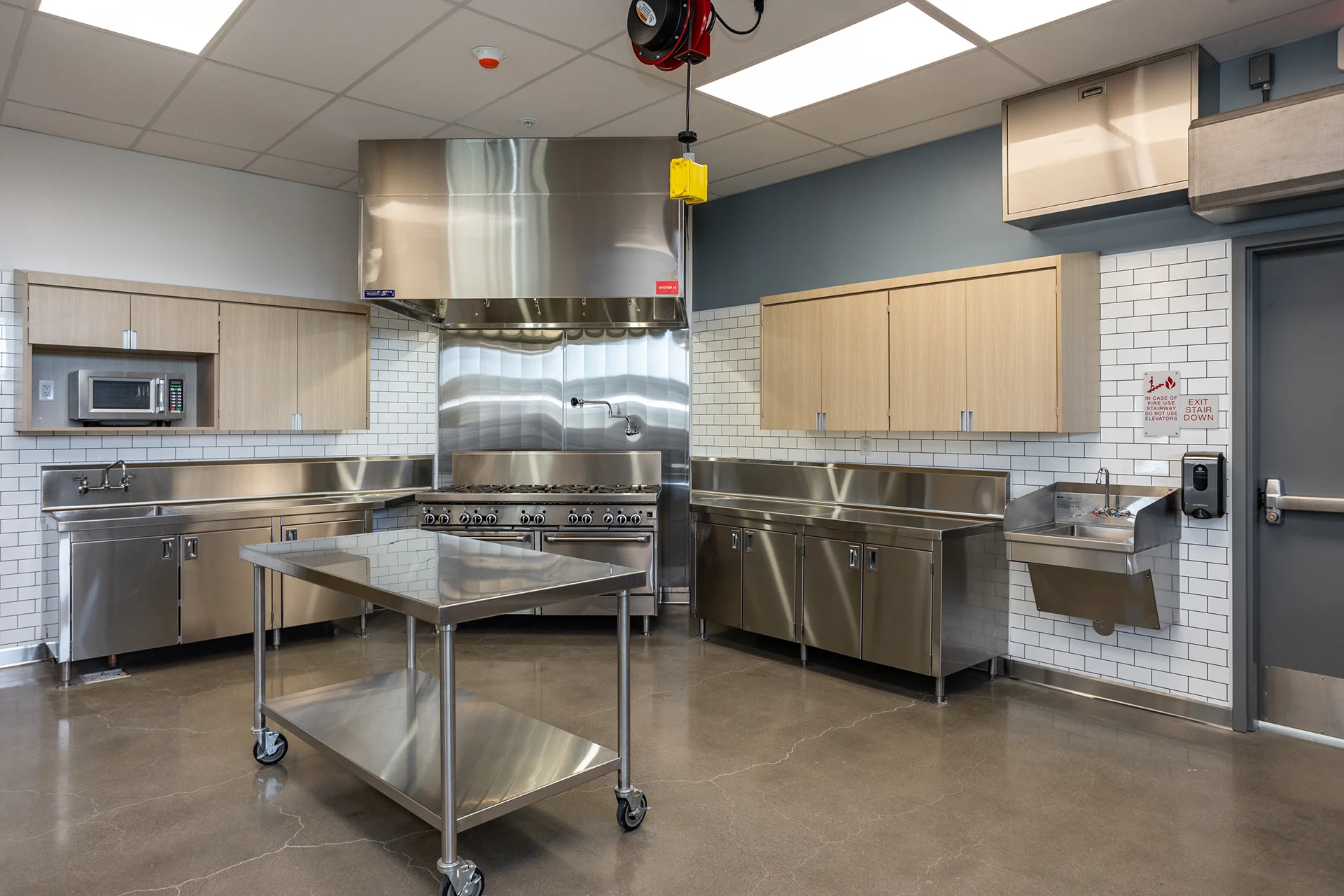 Institutional teaching kitchen layout showcasing a rolling stainless steel work table over a sealed concrete floor, flanked by industrial gas ranges under a large ventilation hood, wood-grain cabinetry, and white subway tile backsplashes.