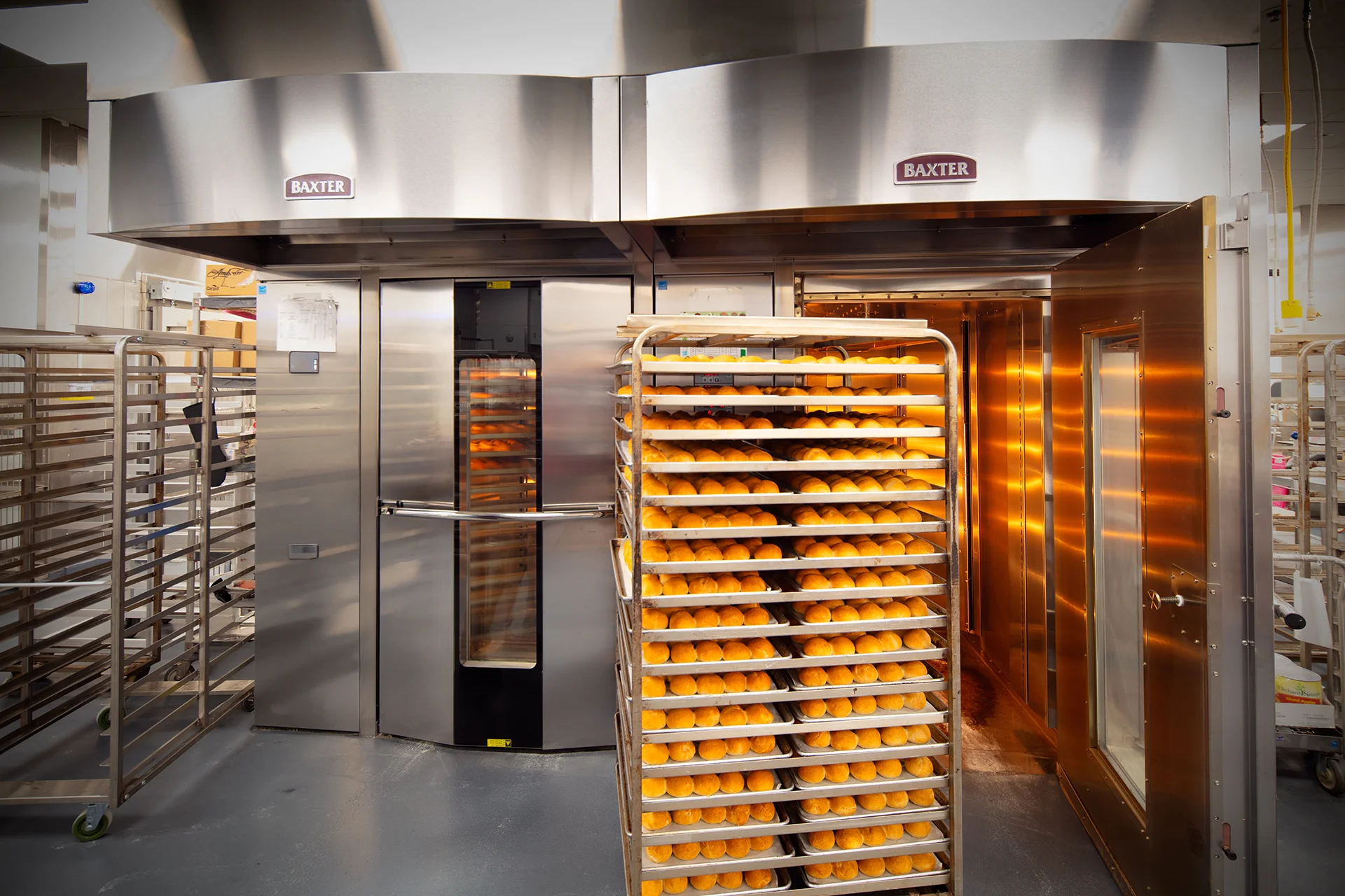 High volume commercial bakery setup featuring twin Baxter stainless steel rotating roll-in rack ovens with a stainless steel exhaust hood, showing a fully loaded mobile cooling rack packed with freshly baked bread rolls.