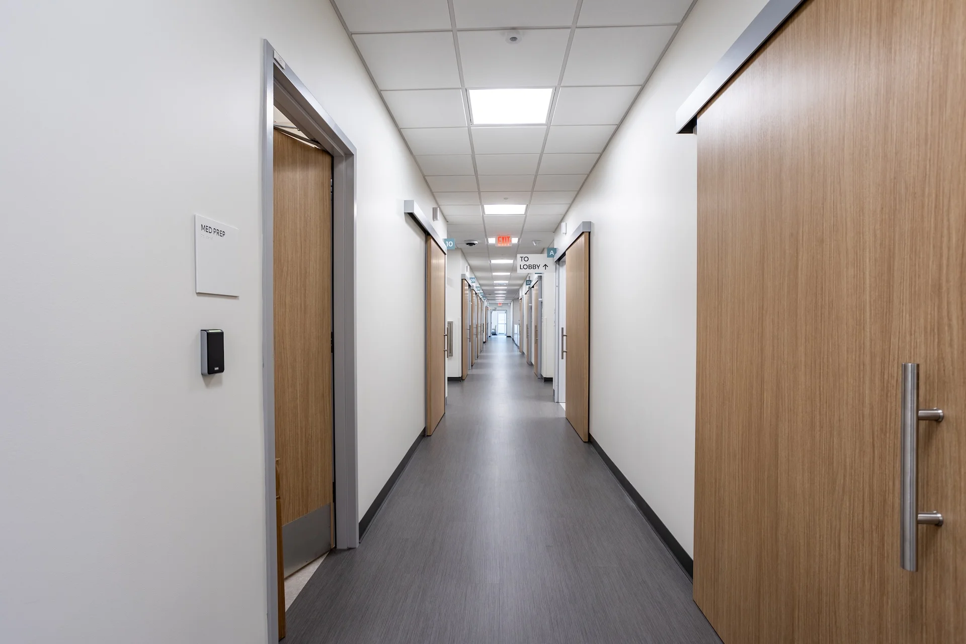 Long interior view of a commercial healthcare facility hallway, utilizing wood-look vinyl plank flooring, white walls, flush acoustic ceiling tiles, clear wayfinding signage, and automated wood veneer sliding pocket doors designed for optimized ADA clearance.