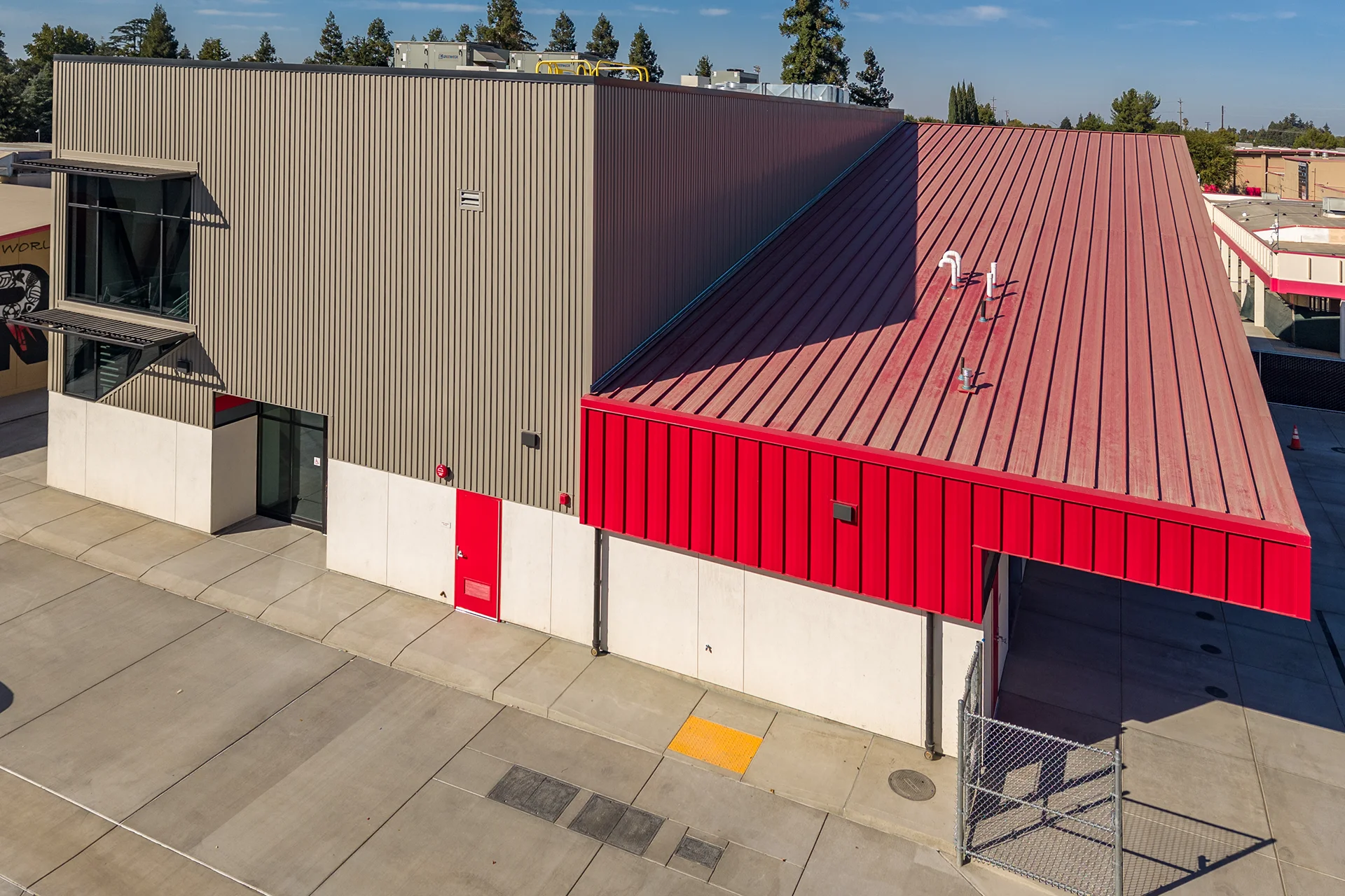 High-angle architectural view of a modern industrial building featuring dual-tone corrugated metal wall panels, a bold red sloped standing-seam roof system, white concrete foundation walls, and concrete sidewalks equipped with ADA tactile warning tiles.