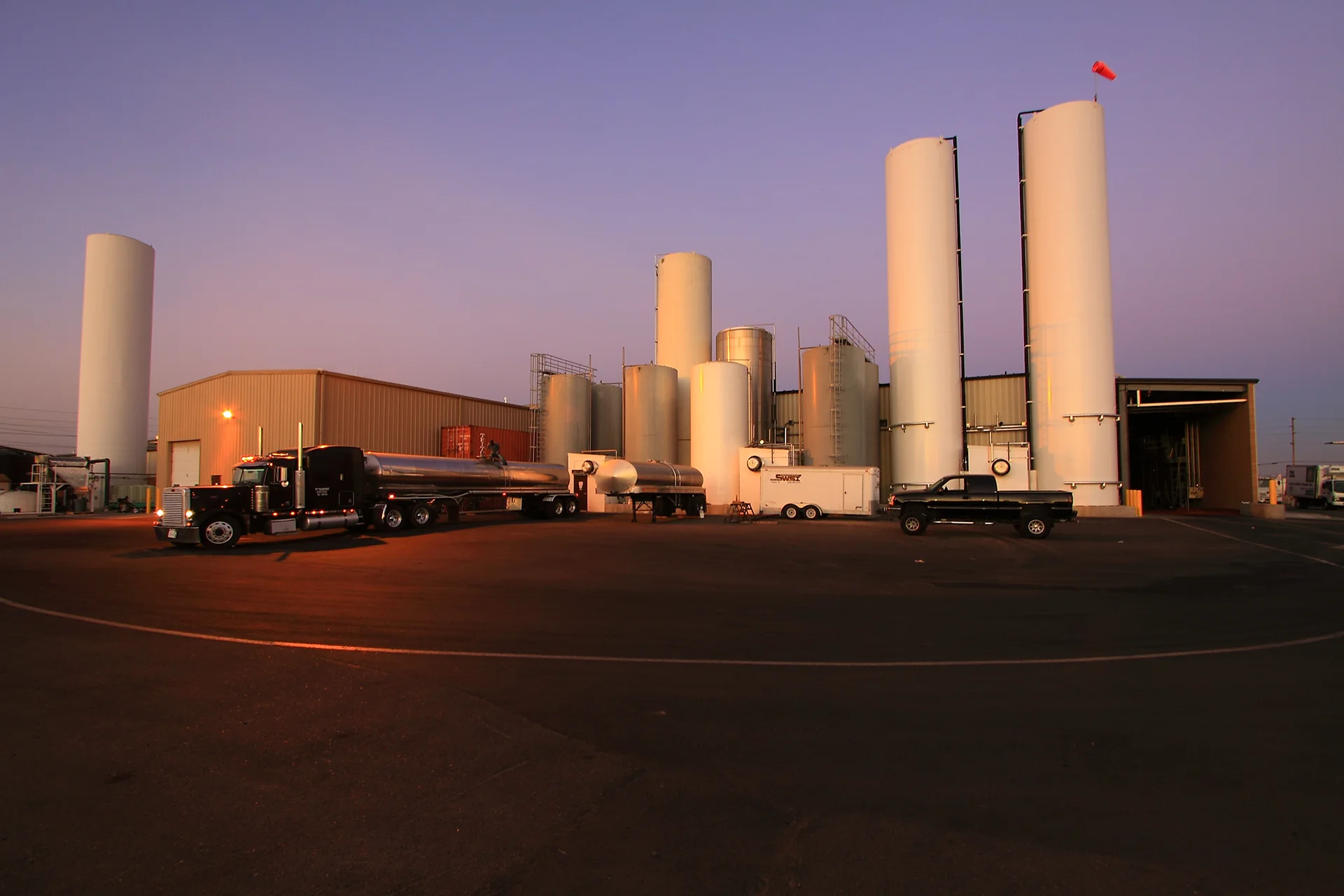 Exterior wide view of an industrial food processing facility at dusk, showcasing an array of vertical white and stainless steel product storage silos, a corrugated metal warehouse building, a parked semi-truck with a stainless steel liquid tanker trailer, and a support vehicle on a clean asphalt lot.