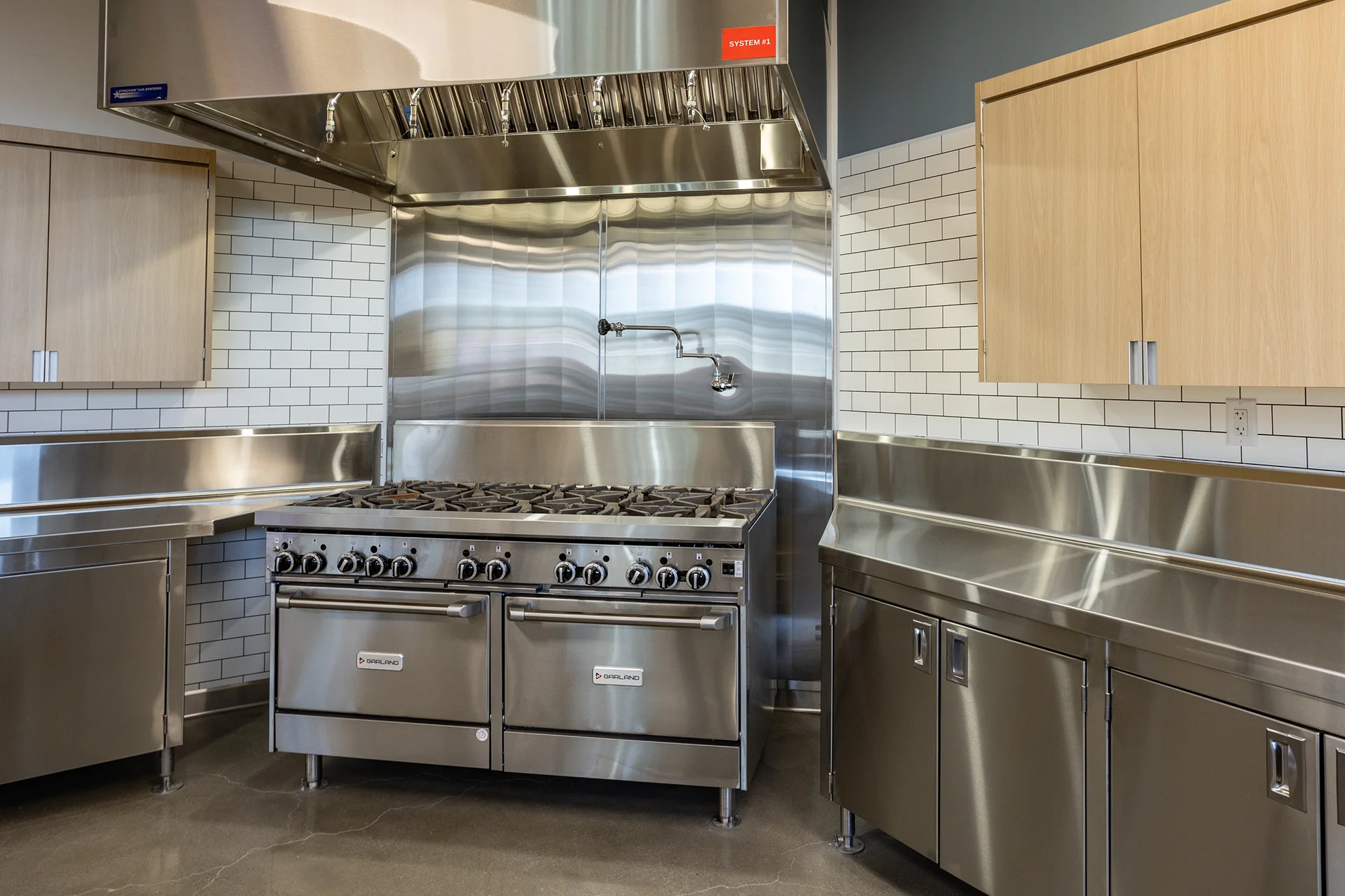 Angled perspective of an institutional cooking station featuring dual commercial ovens, integrated marine-edge stainless counters, custom light-wood overhead cupboards, and clean utility outlets.