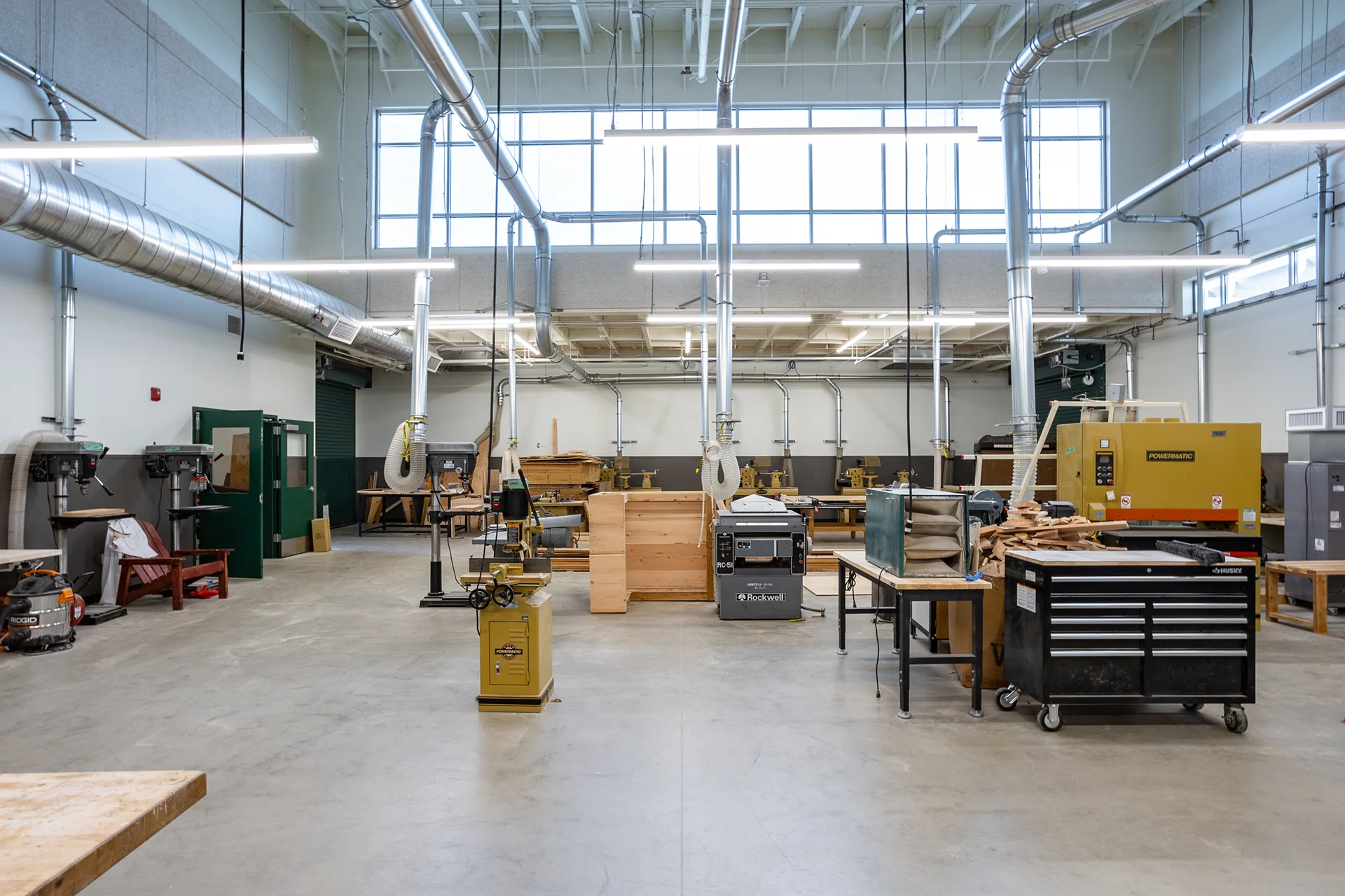 Spacious interior of a high school vocational woodworking facility in Manteca, California, displaying professional-grade machinery including drill presses, a Rockwell planer, and a Powermatic sander, integrated with overhead source-extraction ventilation ductwork, high window bands, and concrete flooring.
