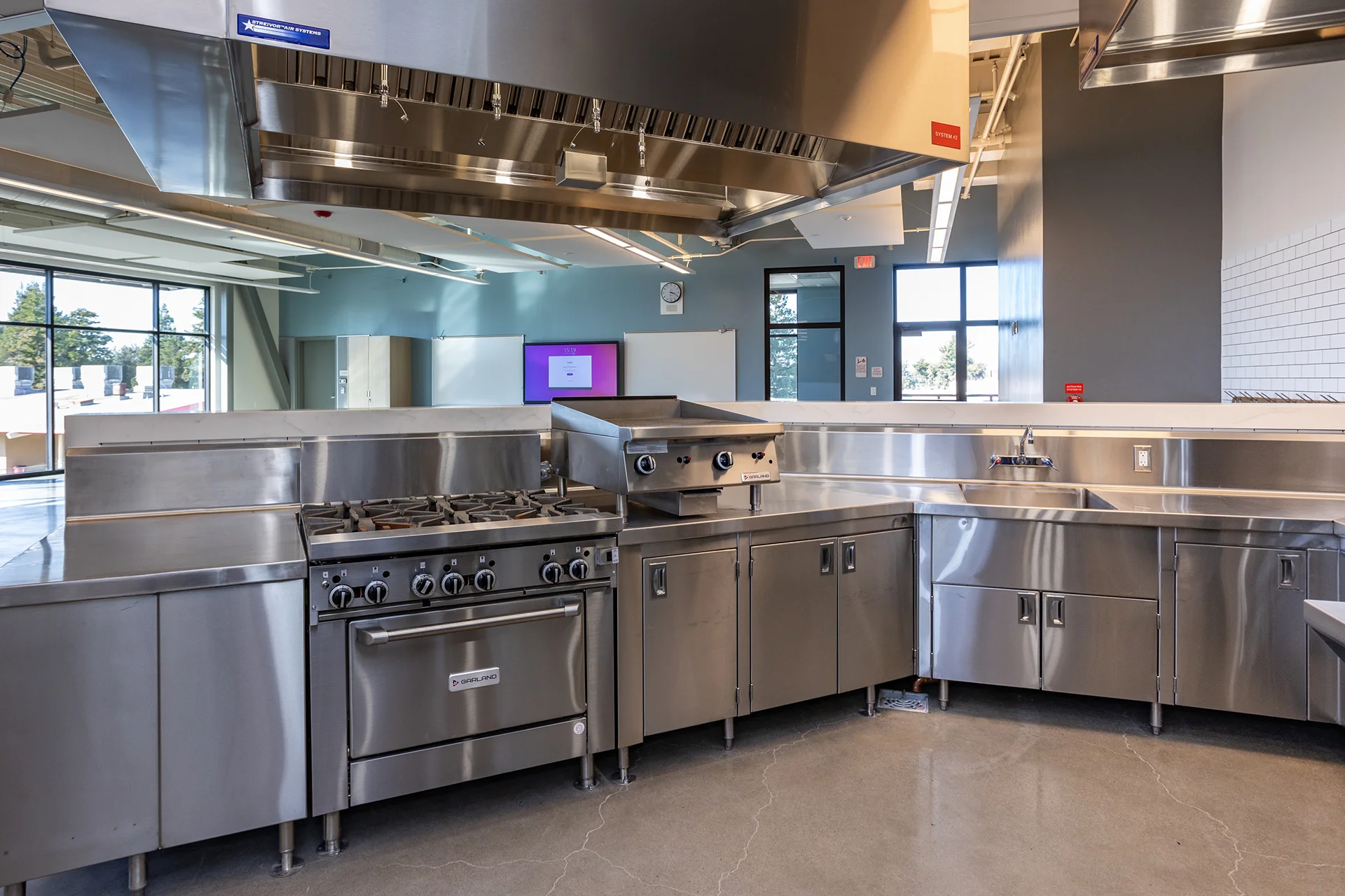 Angled view of a stainless steel culinary station featuring an eight-burner gas range, a counter-mounted flat-top griddle unit, and an integrated handwashing sink, completely covered by a professional exhaust canopy with commercial baffle filters in a modern training facility.