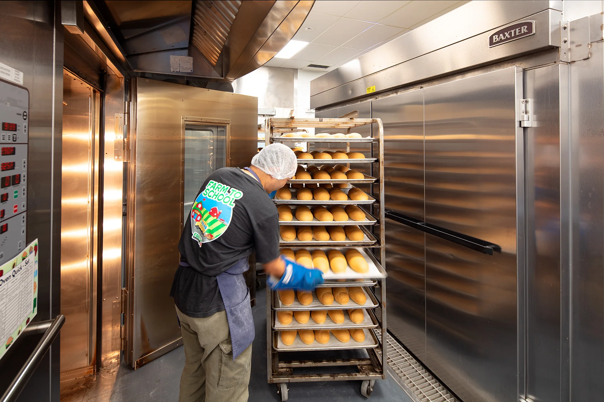 Commercial bakery production view of a worker loading trays of fresh bread loaves onto a heavy-duty stainless steel roll-in cooling rack next to a Baxter industrial rotating rack oven.