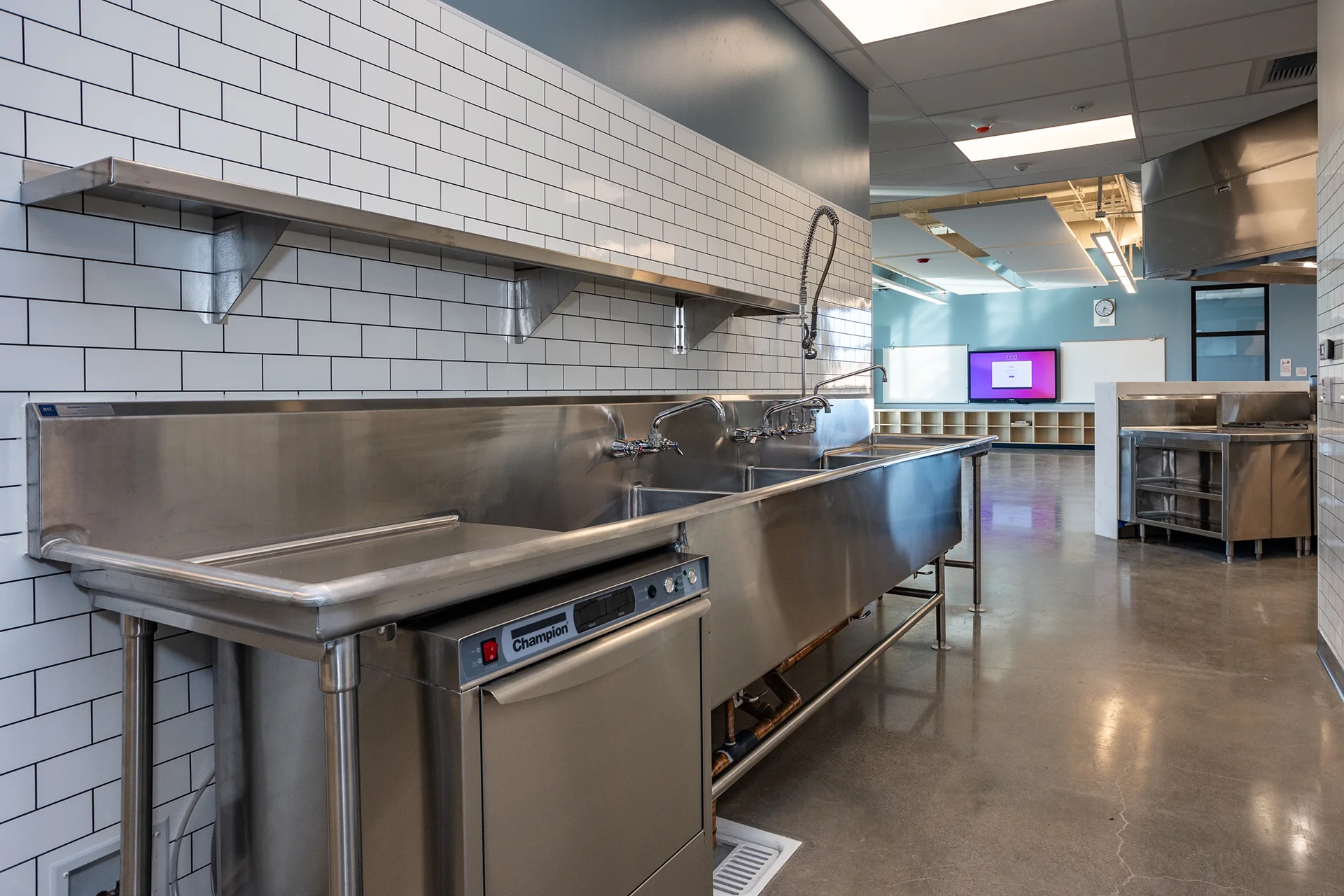 Deep multi-compartment stainless steel wash trough fitted with commercial pre-rinse sprayer faucets, an under-counter automatic commercial dishwasher, an overhead metal storage shelf, and high-gloss white tiled walls.