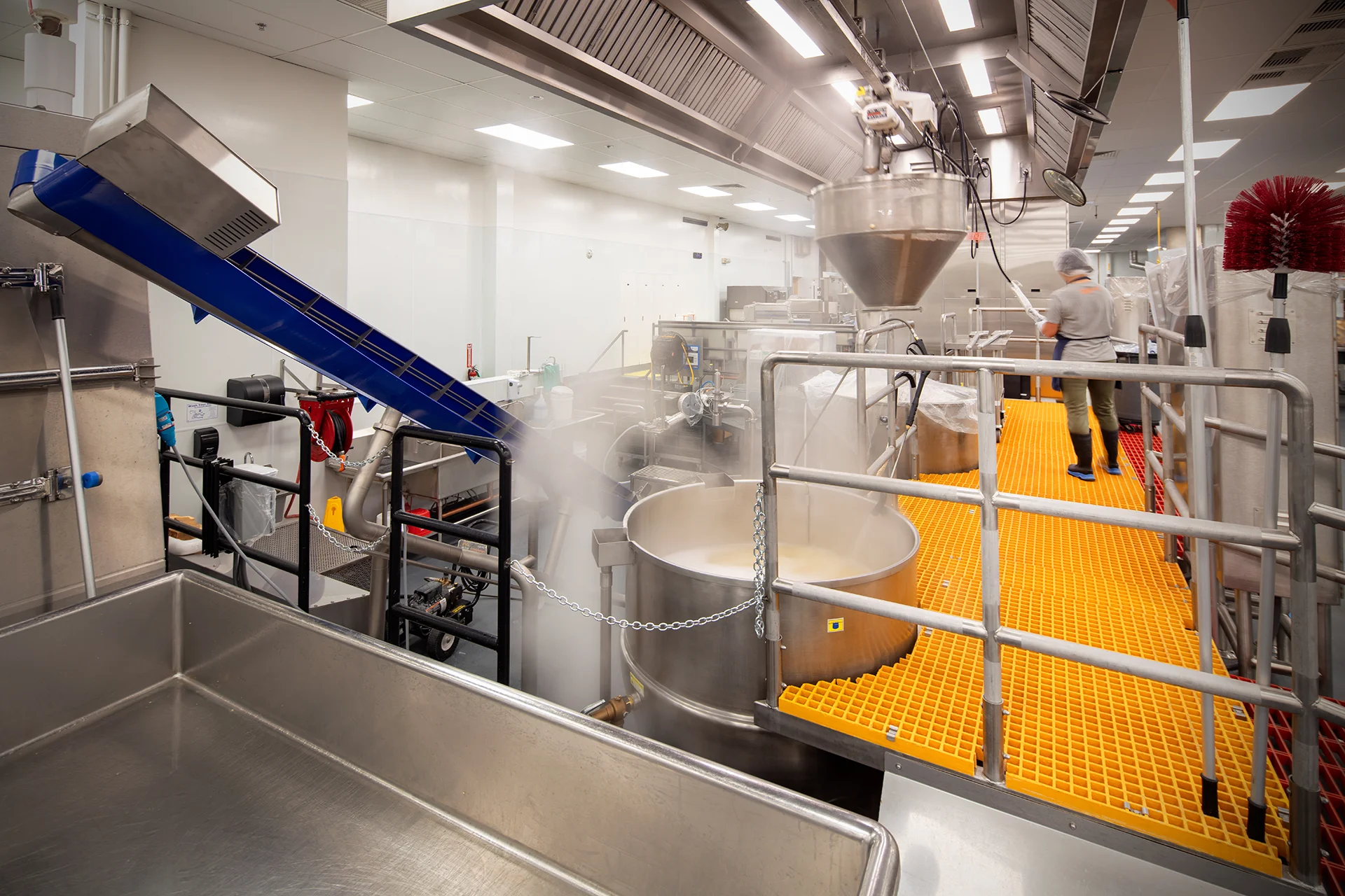 Industrial food production line featuring a large stainless steel steam-jacketed kettle, an integrated inclined blue belt conveyor system, safety handrails, and yellow fiberglass grating platforms engineered for commercial facility code compliance in Elk Grove, California.