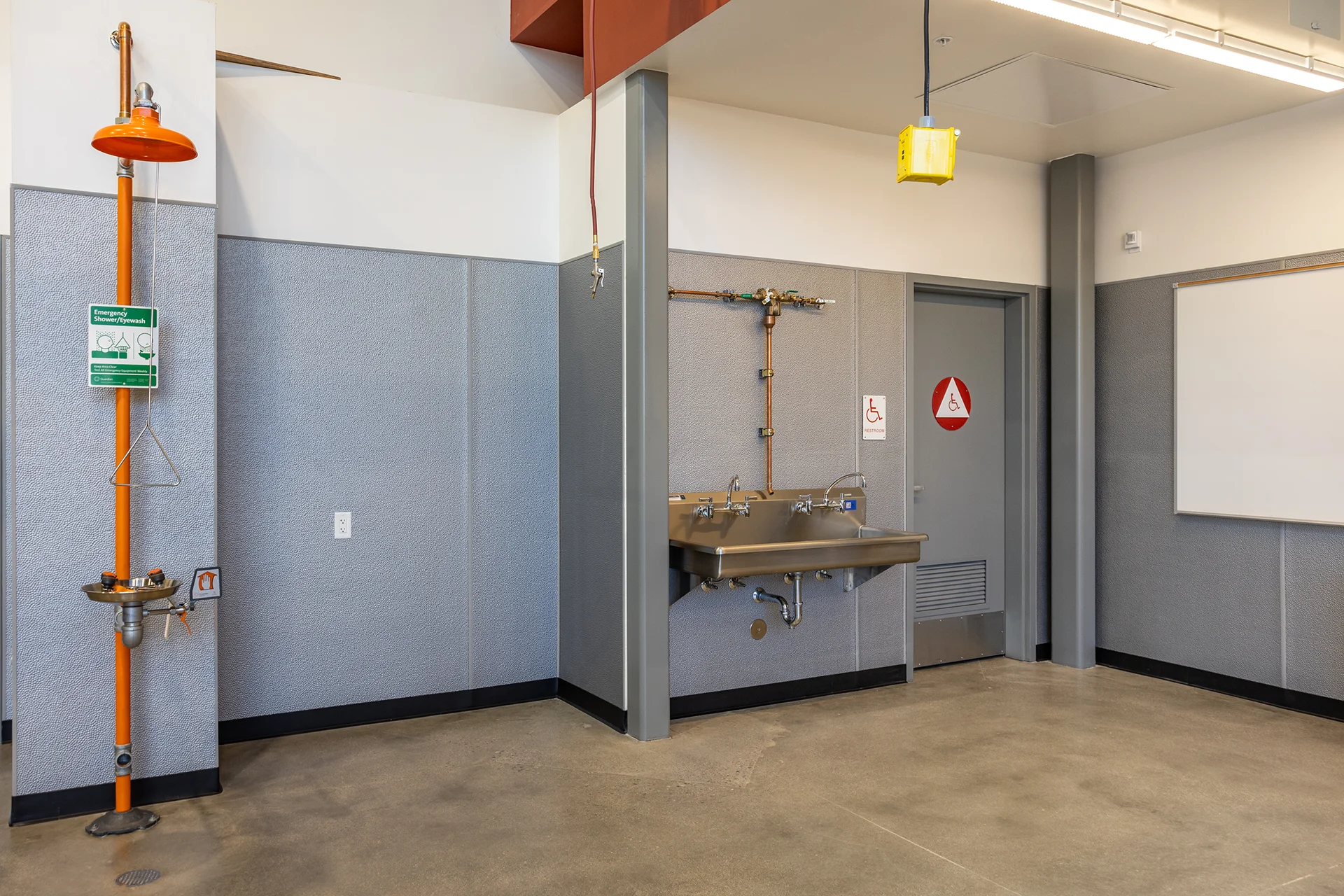Closer perspective of a wall-hung multi-faucet stainless steel scrub sink with exposed brass traps and plumbing, situated between an industrial safety shower and a gray steel commercial restroom door with tactile ADA signage.