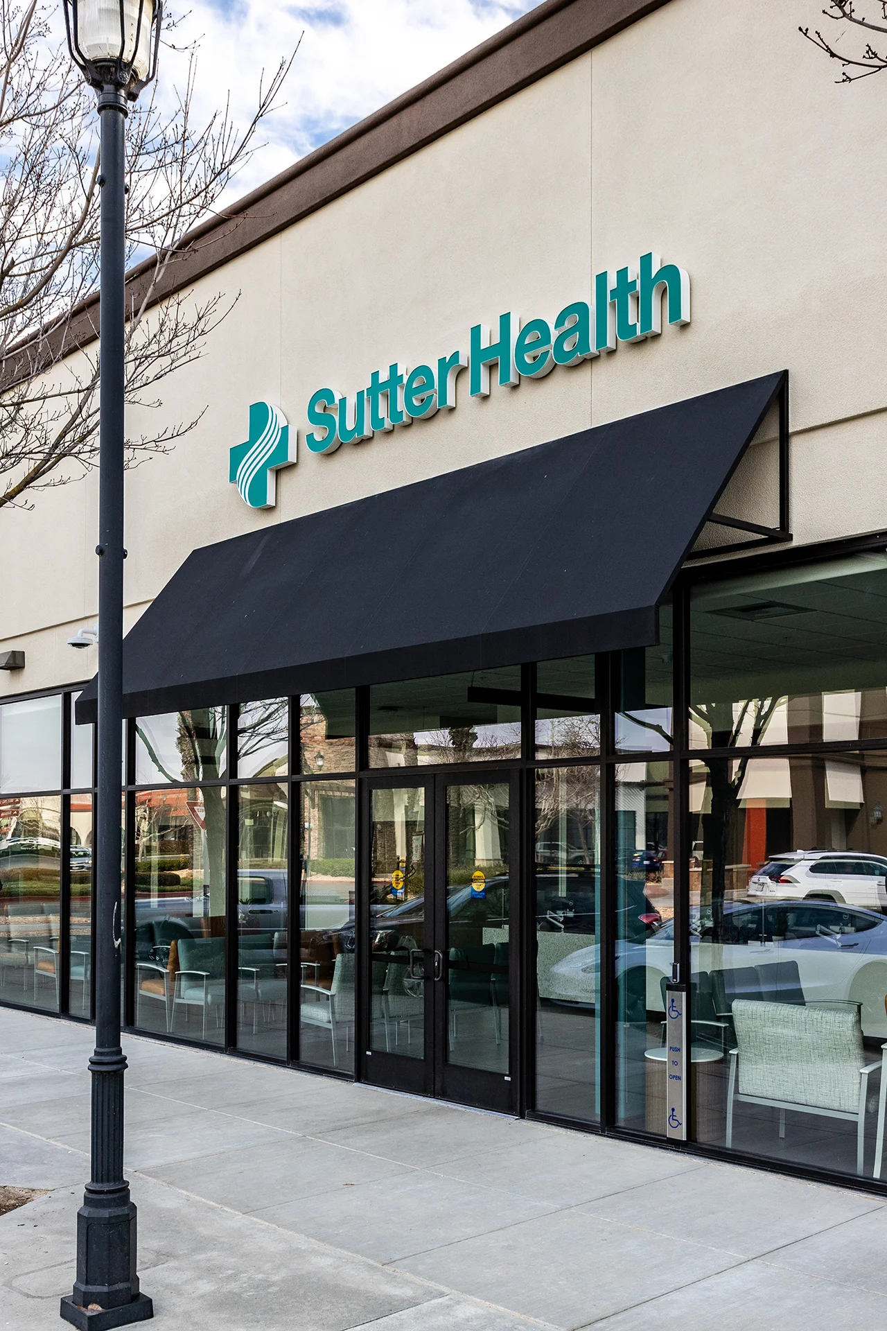 Exterior architectural view of an outpatient clinic entrance featuring dark anodized aluminum storefront window frames, dual glass entry doors with an automated ADA push-plate column, a black canvas wedge awning, and dimensional corporate exterior signage.