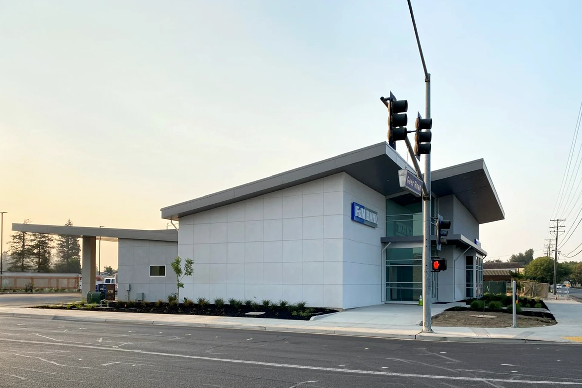 Wide exterior profile shot of the completed commercial construction for F&M Bank at the corner of Geer Road, featuring a dramatic sloped metal roofline, perimeter landscaping beds with dark mulch, and an adjacent drive-up banking canopy structure.