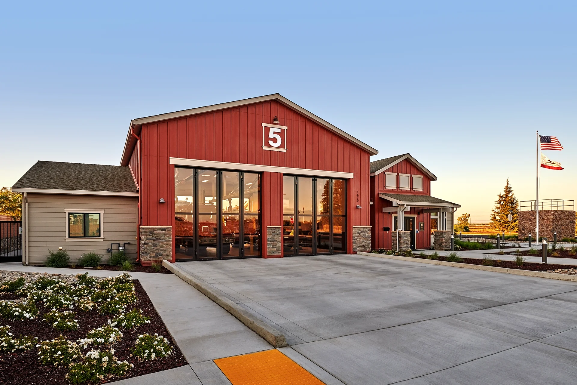 Wide exterior shot of the fully completed civic construction project, showing closed glass bifold garage doors reflecting the sunset sky, clean concrete paths, landscaped beds, and municipal flags flying.