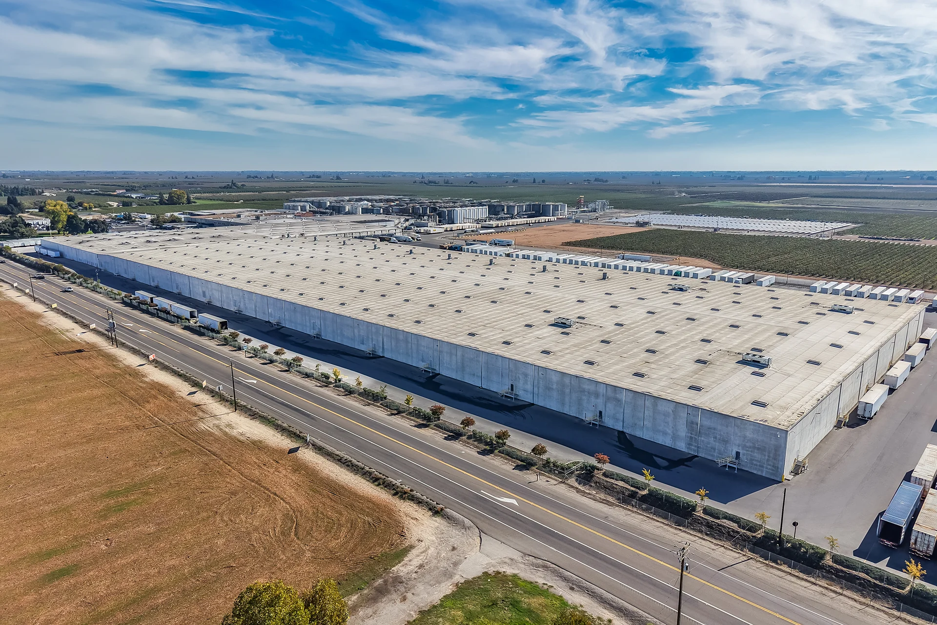 Panoramic high aerial shot of an expansive concrete industrial manufacturing facility, detailing a continuous low-slope corrugated metal roof assembly, an adjacent multi-lane highway, and an extensive background matrix of stainless steel bulk fluid storage silos and processing infrastructure under a clear sky.