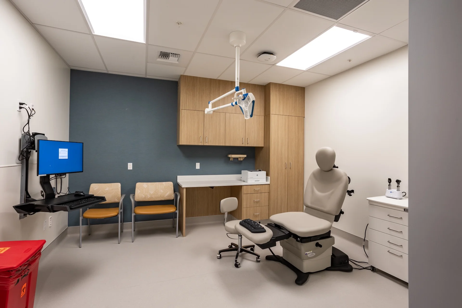 Premium medical exam space fitted with light oak laminate overhead cabinets and full-height storage wardrobes, an articulating overhead examination light, a modular clinician stool, standard diagnostic instruments on a mobile white cart, and an articulating gray upholstered examination table.