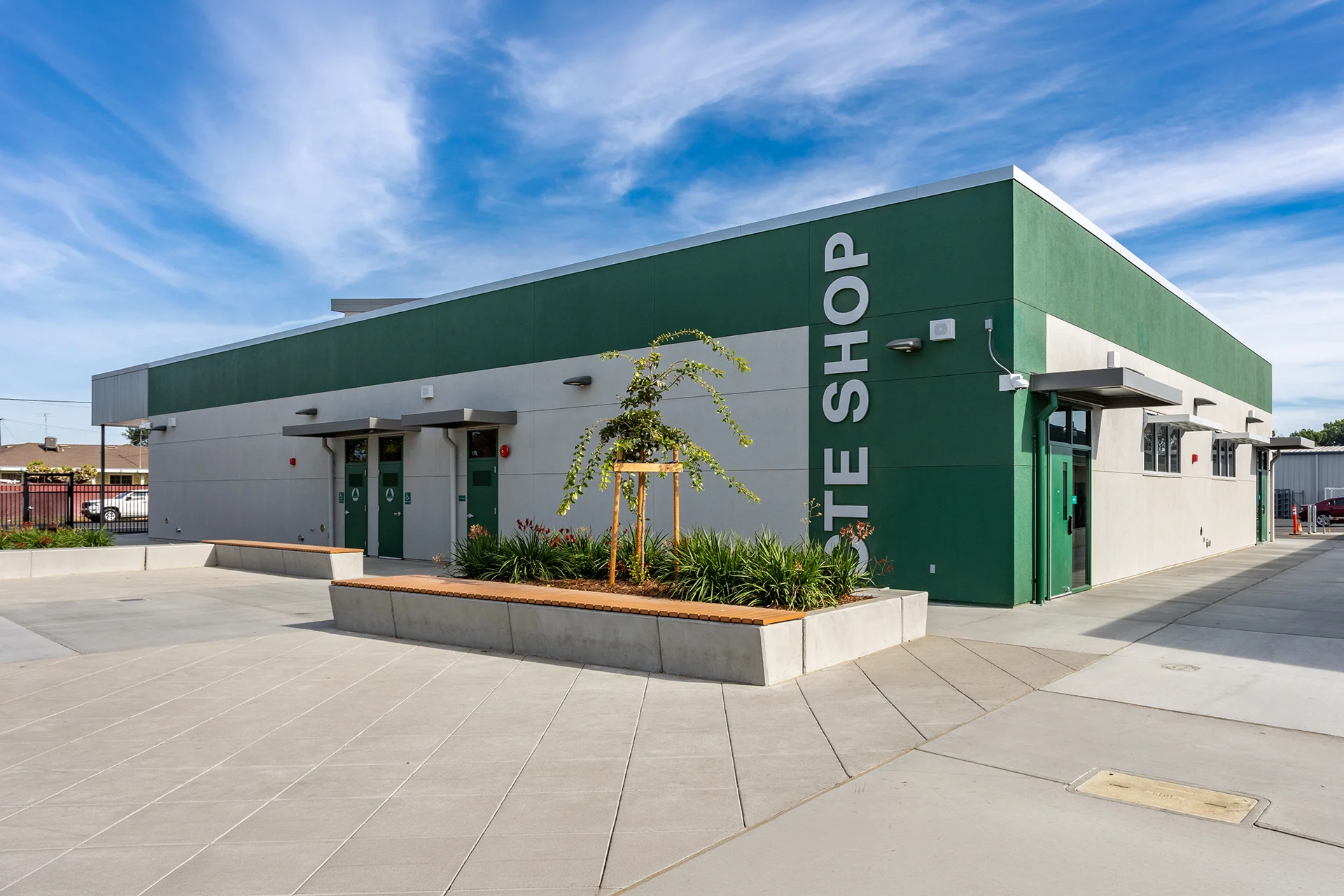 A modern institutional workshop facility labeled Site Shop, featuring a prominent dark green and gray stucco exterior, green commercial exit doors, steel awnings, geometric concrete paver walkways, and a integrated concrete bench planter with wood slat seating.