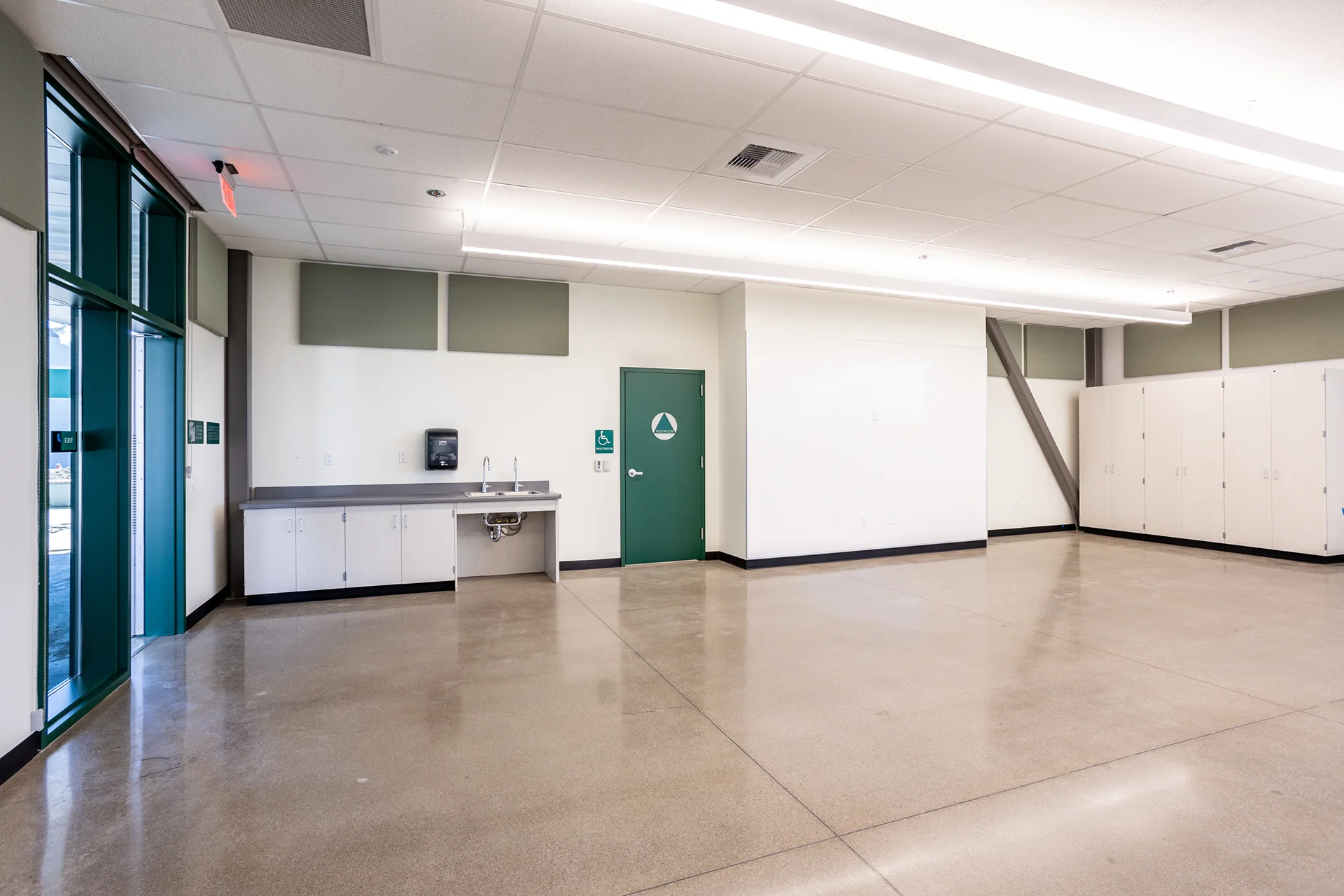 Multi-functional instructional room interior in Manteca, California, showcasing a sleek utility alcove with dual stainless steel commercial faucets, a green bathroom door with a geometric ADA emblem, clean acoustic panels, and high gloss architectural flooring.