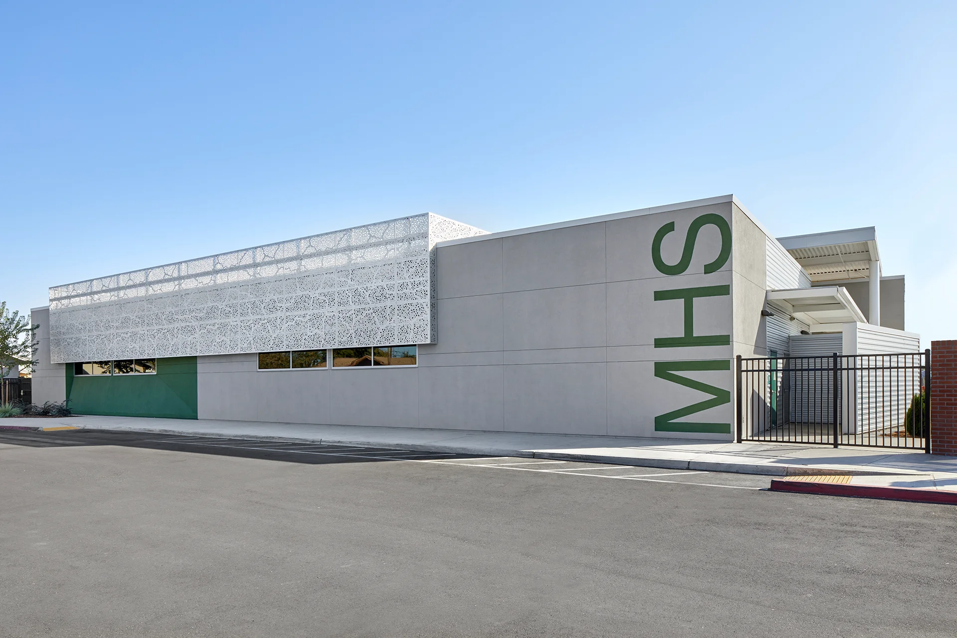 Ground-level view of an educational building in San Joaquin County, showing a minimalist grey stucco exterior wall detailed with massive dark green vertical vinyl lettering, an integrated geometric metal screening partition, and a black wrought-iron security fence.