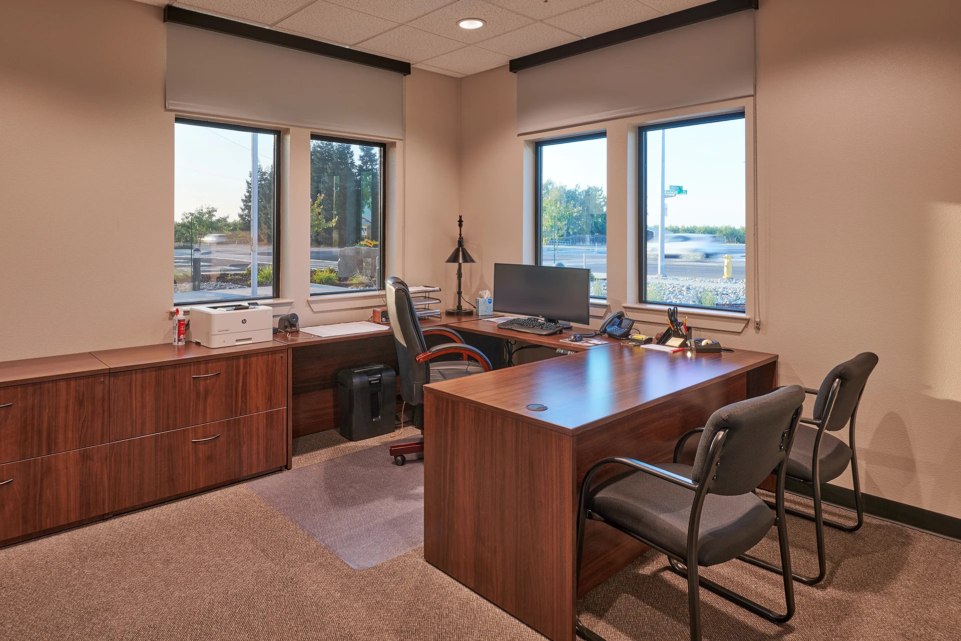 Well-lit administrative office featuring a wooden executive desk, guest seating, multi-pane windows looking out toward a street corner, and integrated window roller shades.