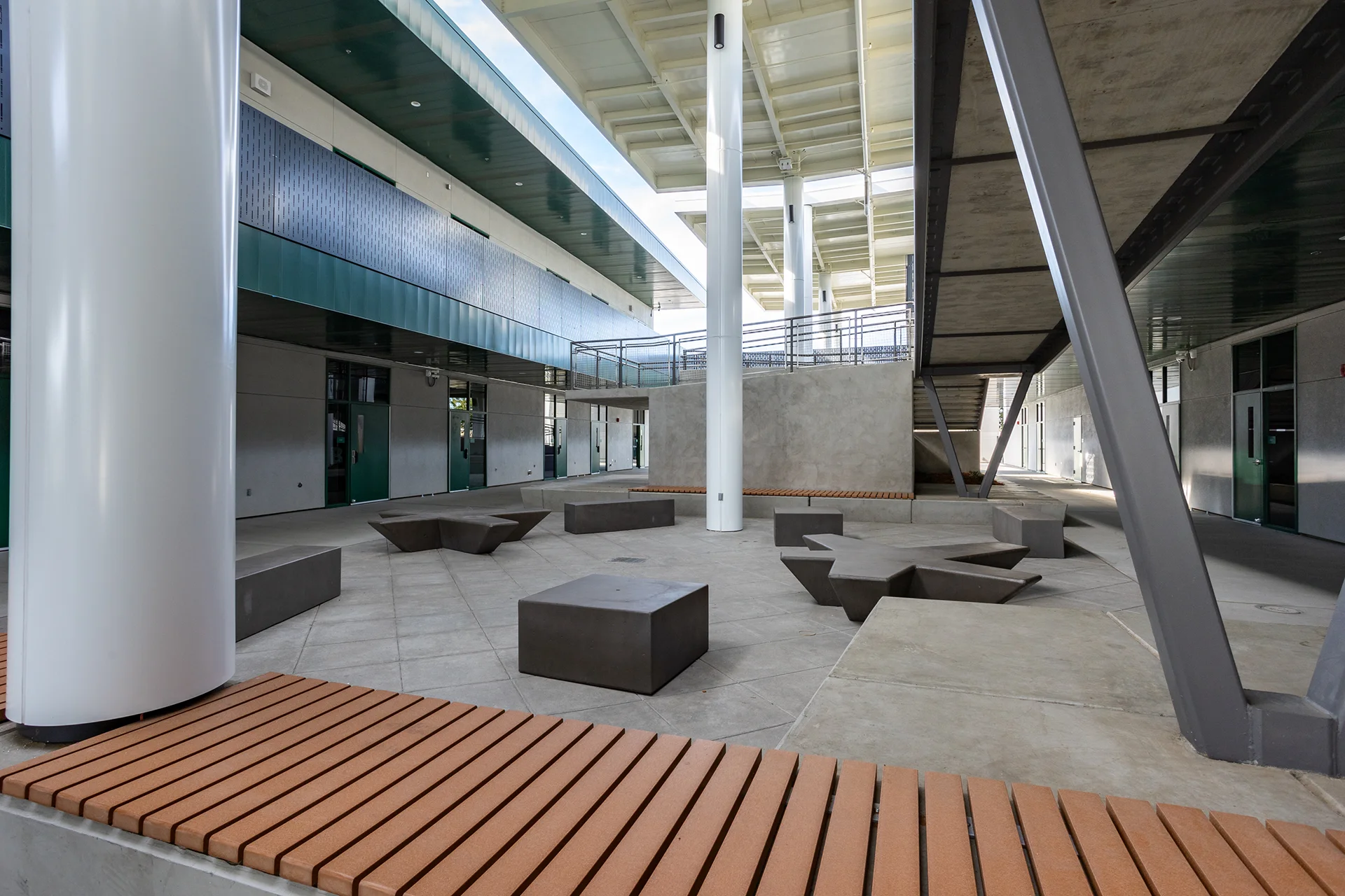 Wide-angle view from a concrete bench with wood slat seating looking into an institutional campus courtyard in San Joaquin County, showcasing multiple star-shaped concrete benches, tall white pillars supporting a high-clearance metal roof canopy, and an elevated concrete walkway with mesh railings.